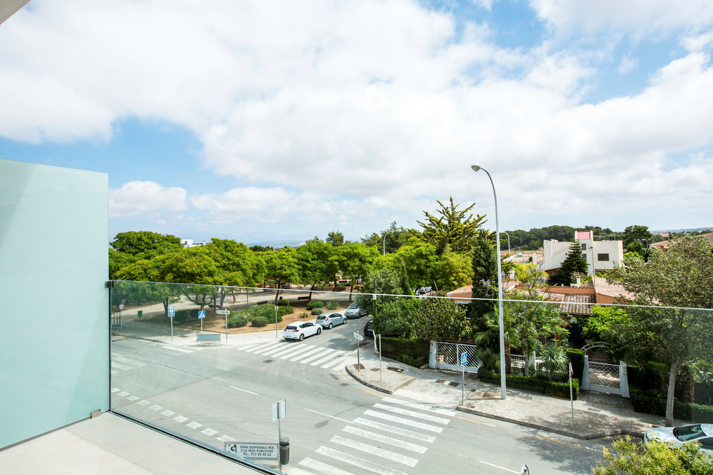 Utsikt fra en balkong med glassrekkverk over en bygate og grøntområder, med park og hus i bakgrunnen. (AI-generert tekst)