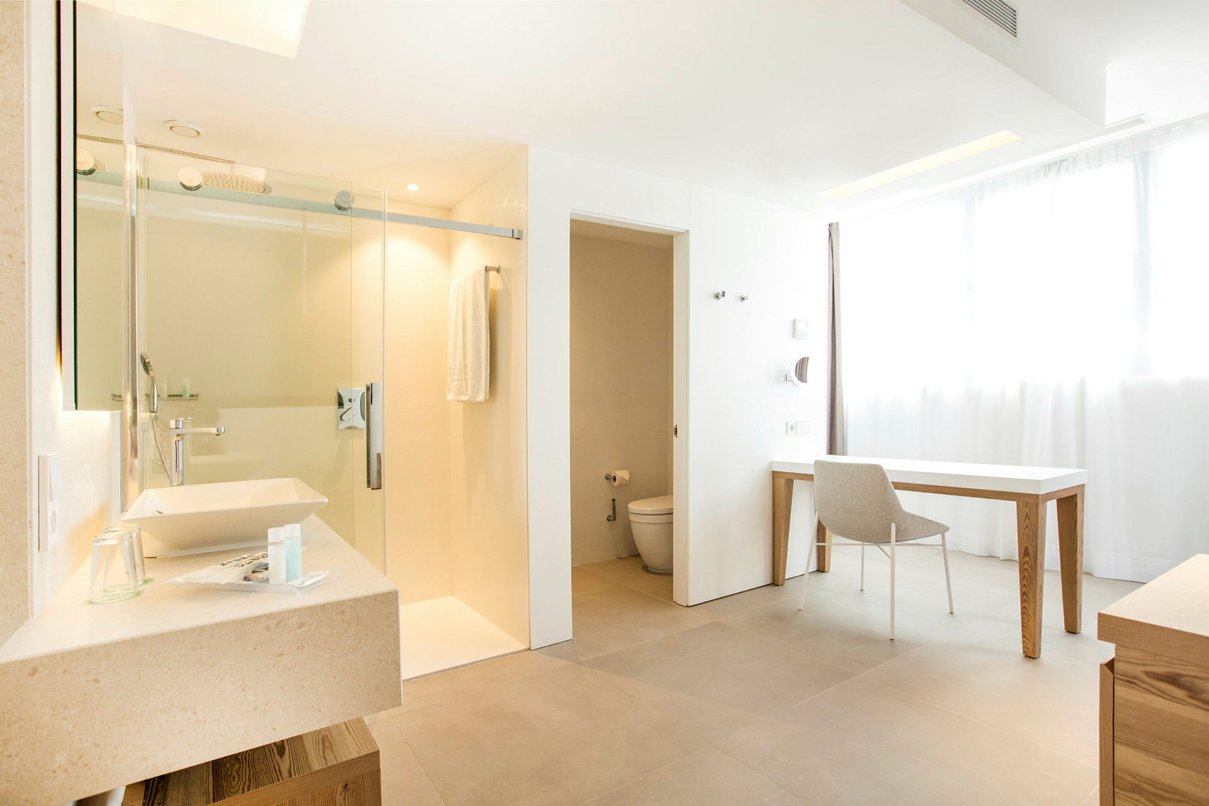 Et moderne hotellrom med baderom i lyse nyanser. Bildet viser en servant, en dusj med glassdør, en toalett og en arbeidsområde med skrivebord og stol. (AI-generert tekst)