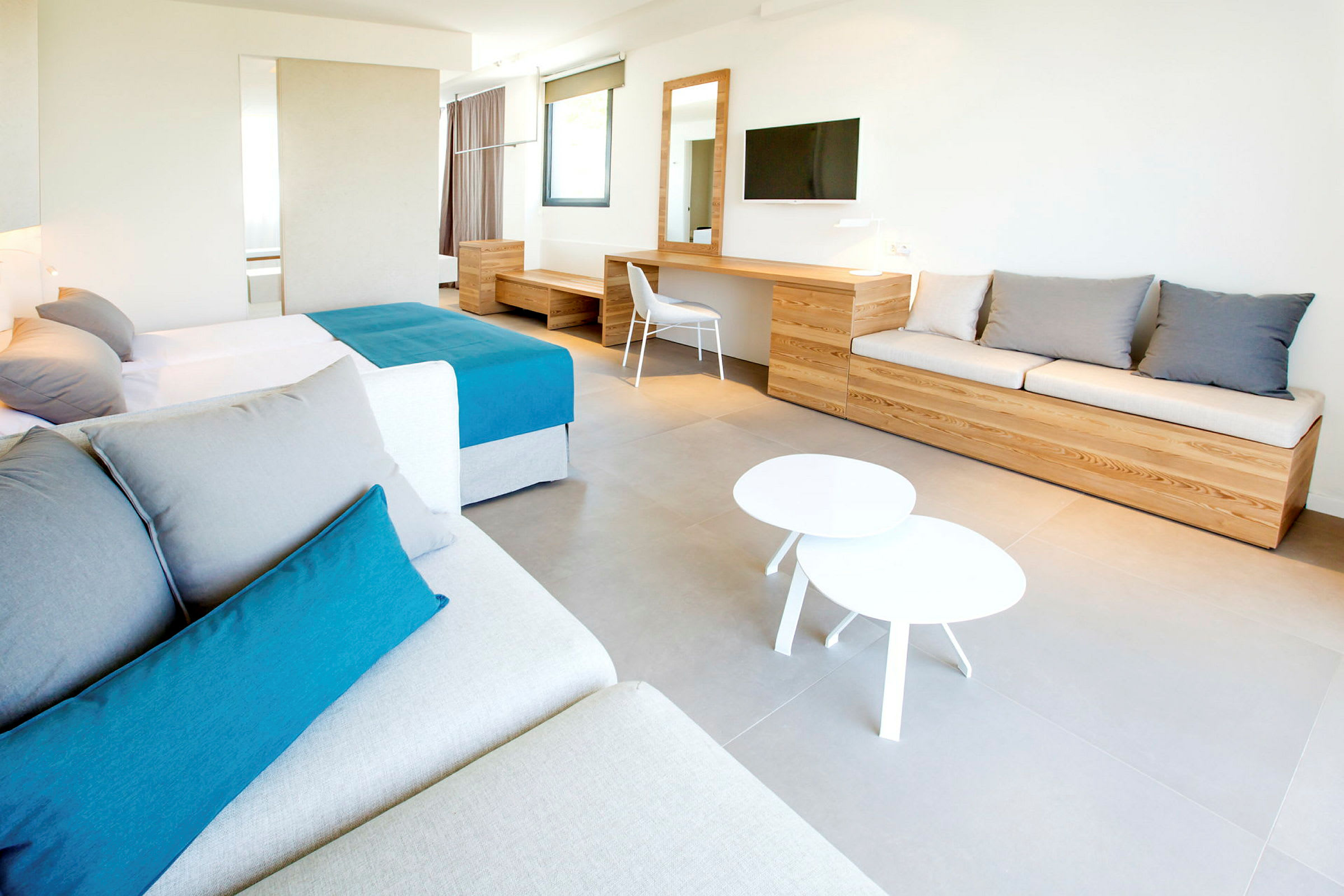 Et moderne hotellrom med en stor seng, en sofa, et skrivebord og en veggmontert TV. Møblene er trefarget, og rommet har en minimalistisk design. (AI-generert tekst)