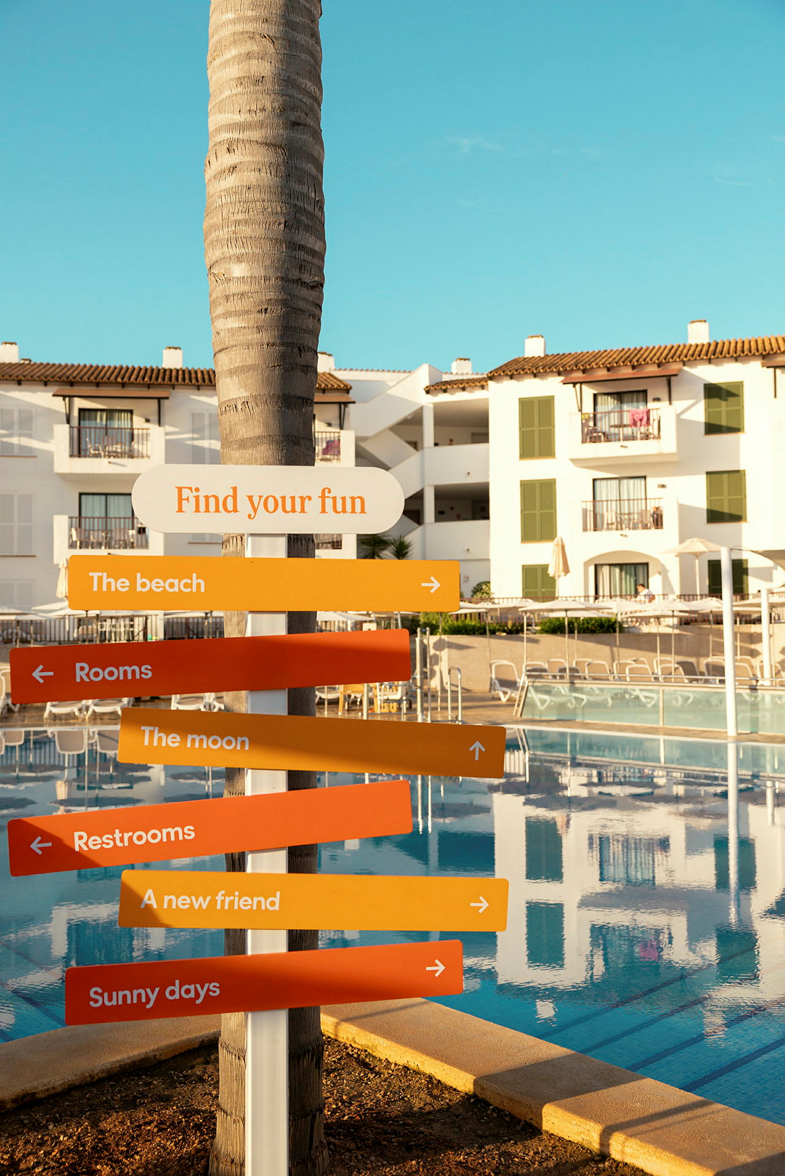 Et skilt med teksten 'Find your fun' og flere fargede piler på en resort med basseng og hvite bygninger. (AI-generert tekst)