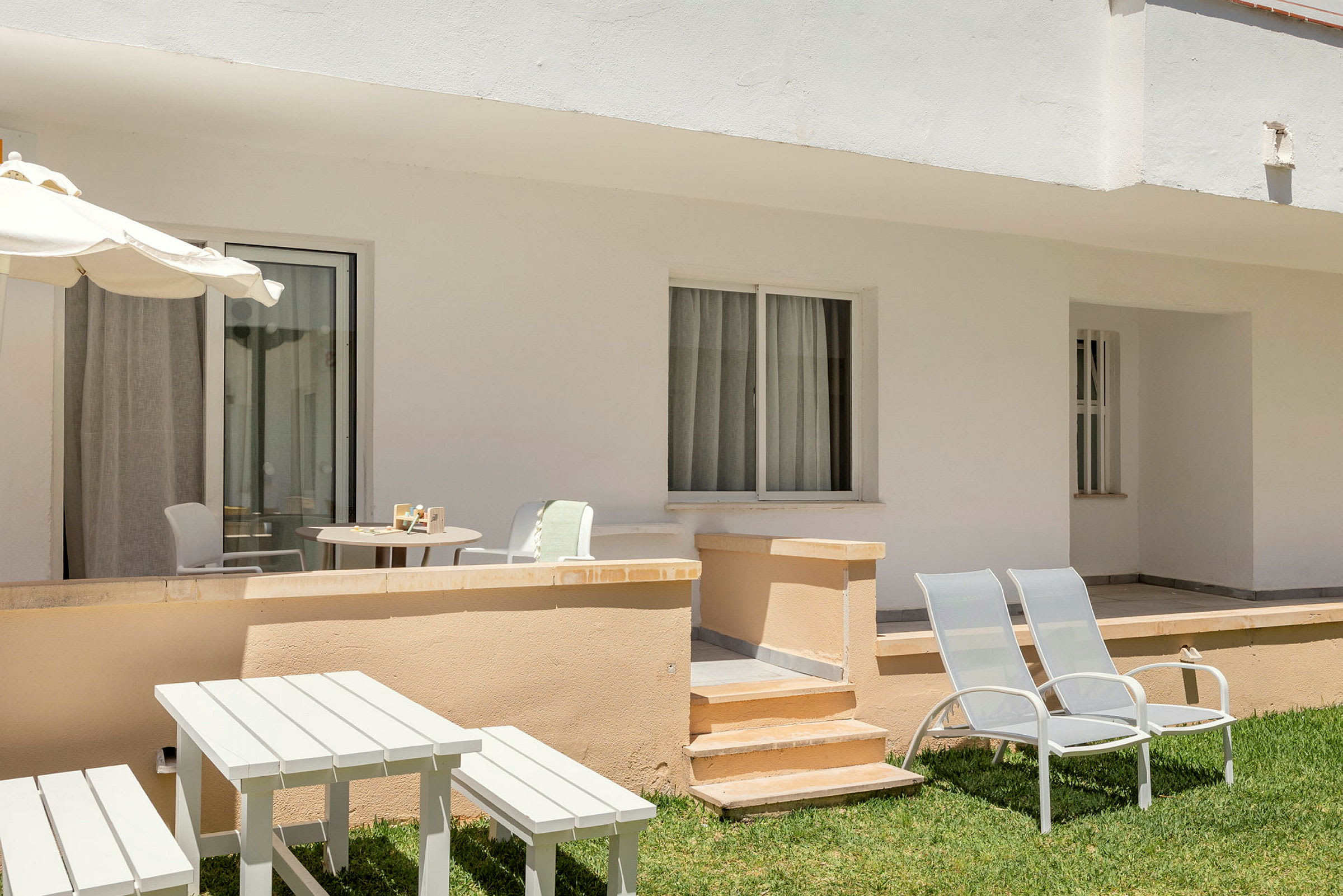 Terrasse med utemøbler og solsenger på en plen utenfor en lys husfasade. (AI-generert tekst)