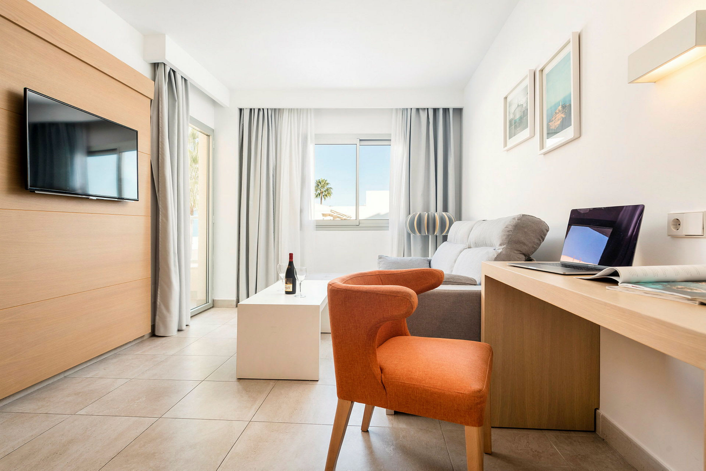 Lyst, moderne hotellrom med sofa, stol, TV, skrivebord og store vinduer. (AI-generert tekst)