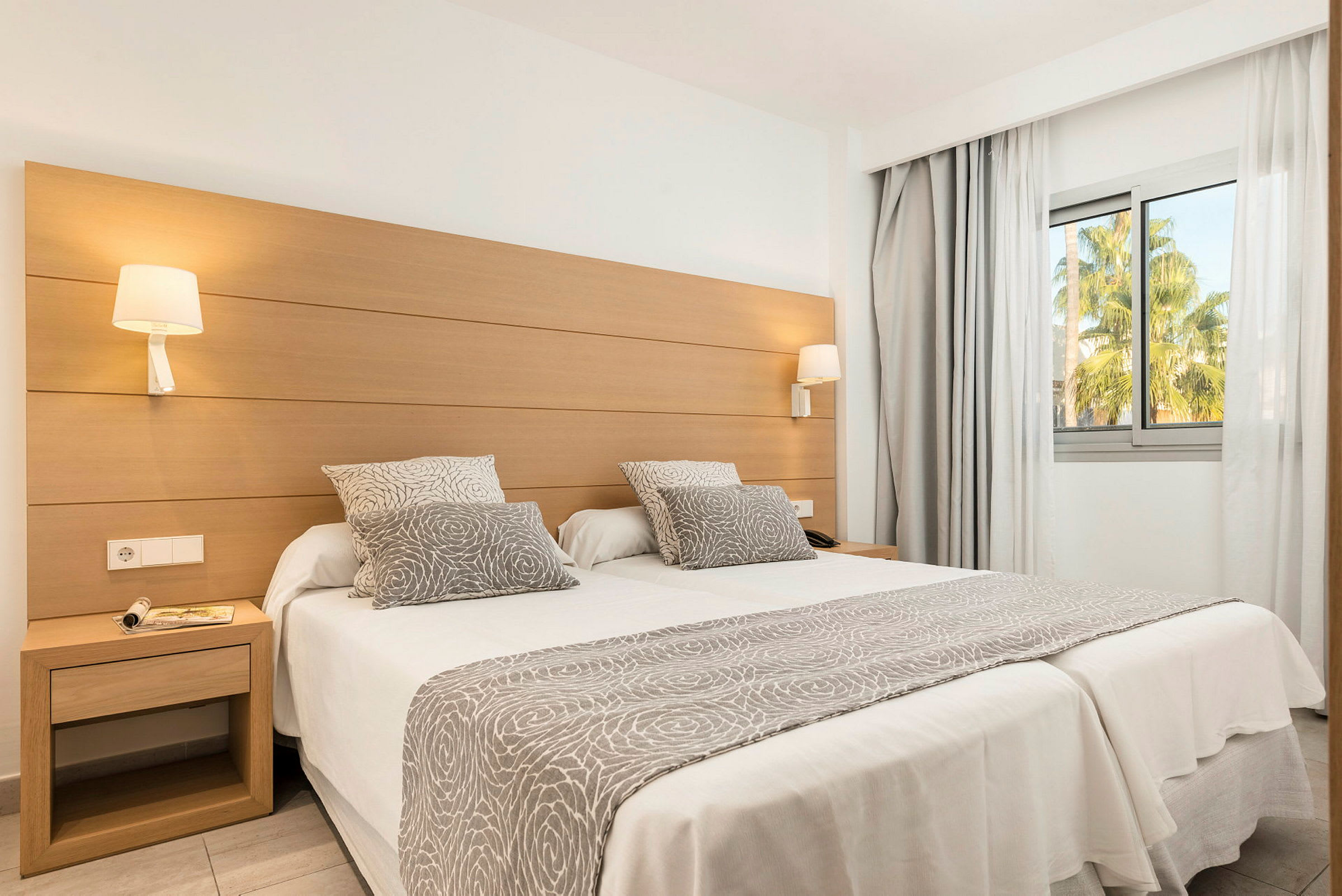Et hotellrom med to enkeltsenger satt sammen, dekorert med gråhvite tekstiler med mønster, og et stort vindu med utsikt mot grøntområder. (AI-generert tekst)