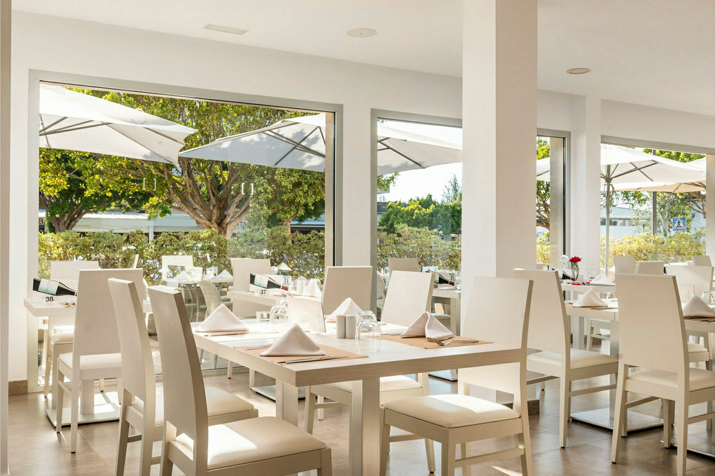 Bilde av en restaurant med hvite stoler og bord, oppsatt med servietter og glass nær store vinduer. (AI-generert tekst)
