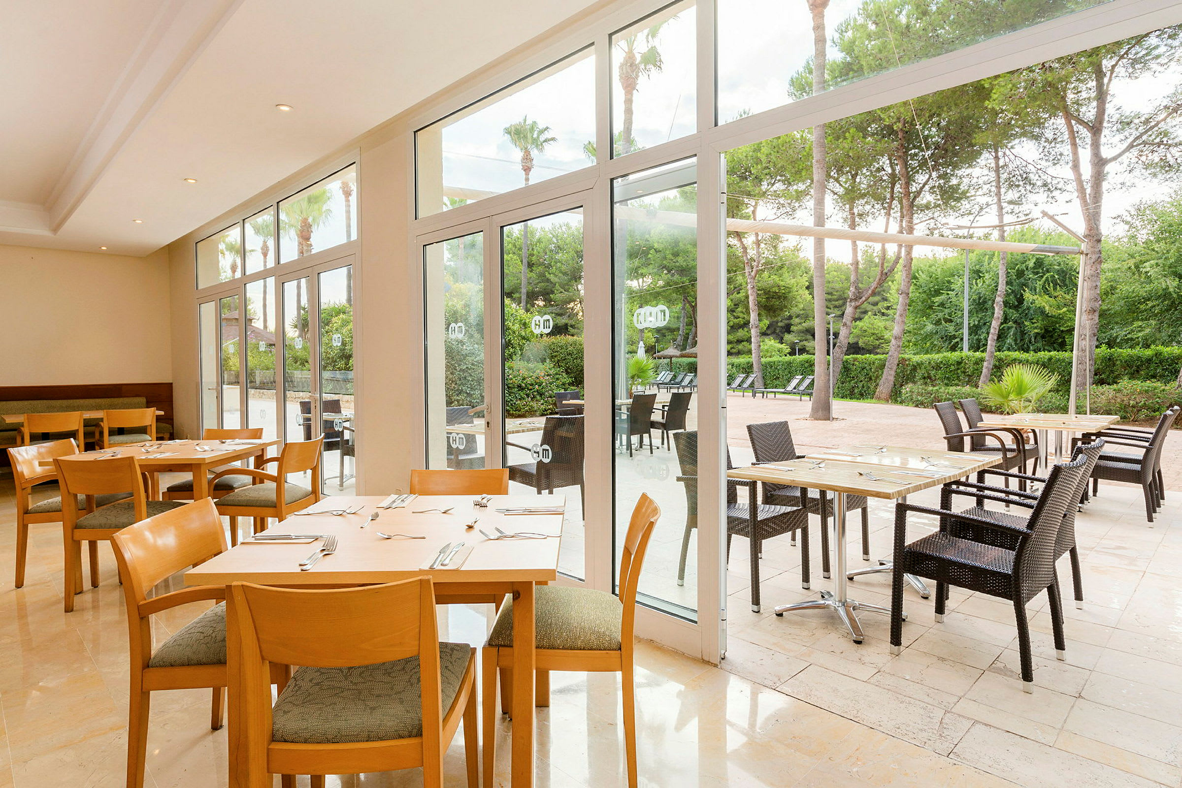 Restaurant med bord og stoler både innendørs og utendørs, med utsikt over hage og trær. (AI-generert tekst)