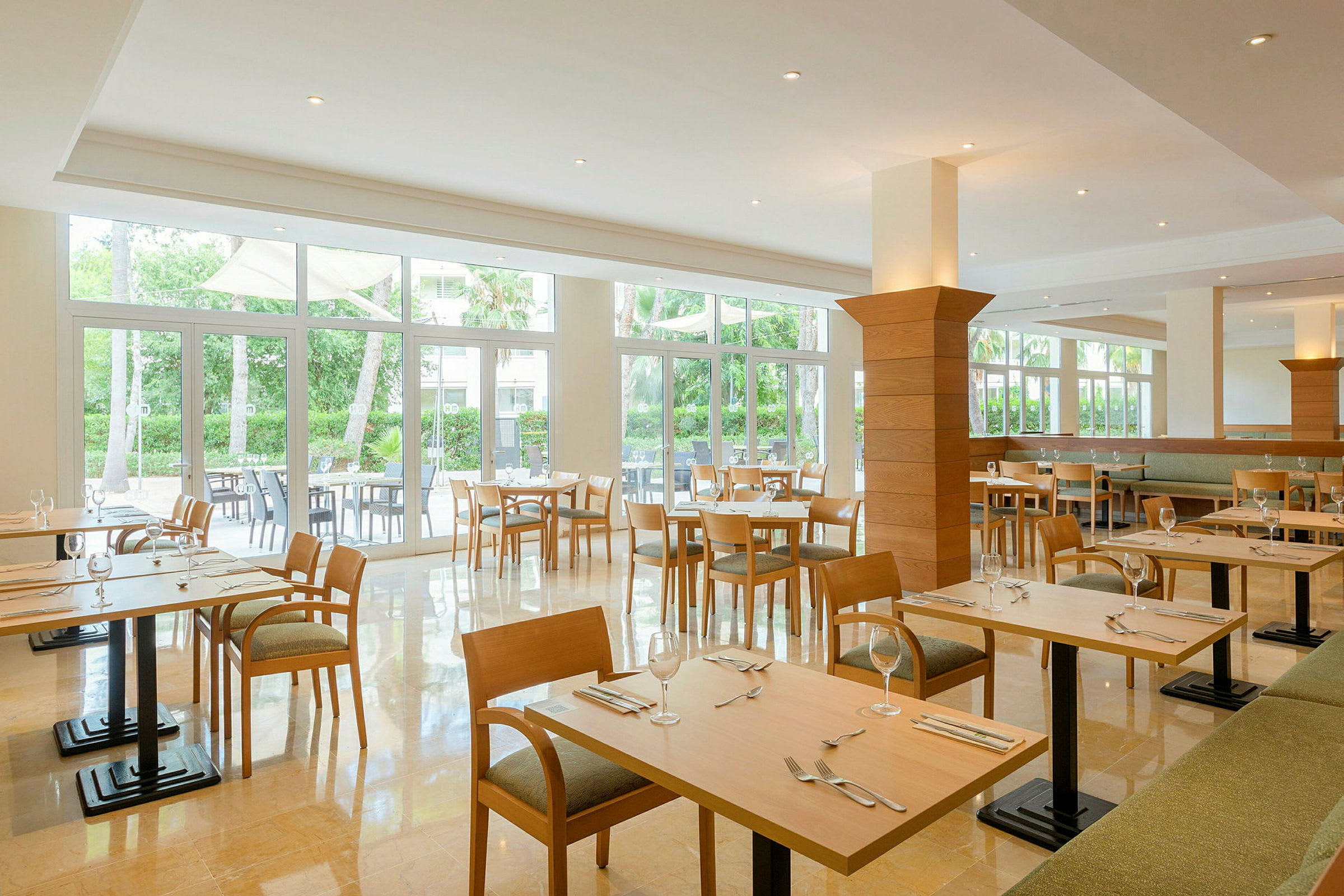 Lys og romslig restaurant med trebord og stoler, omgitt av store vinduer med utsikt over grøntområder. (AI-generert tekst)