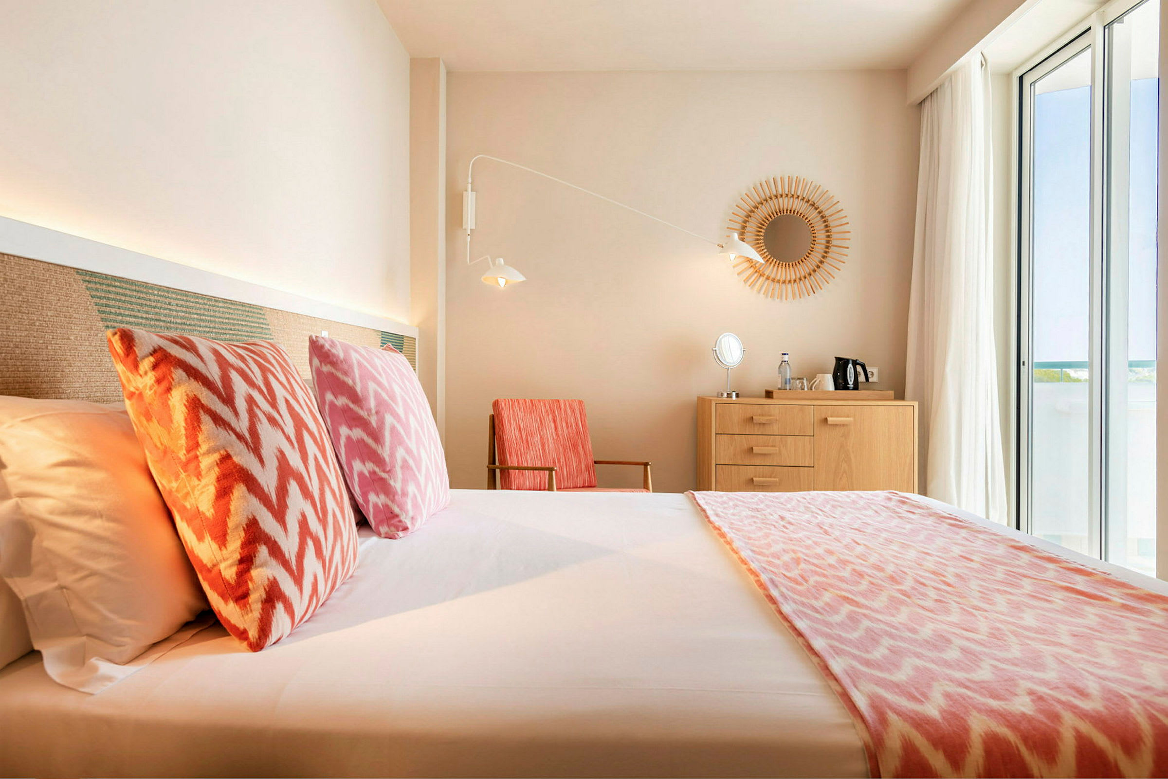 Et hotellrom med en seng, fargerike puter, en trekommode og store vinduer. (AI-generert tekst)