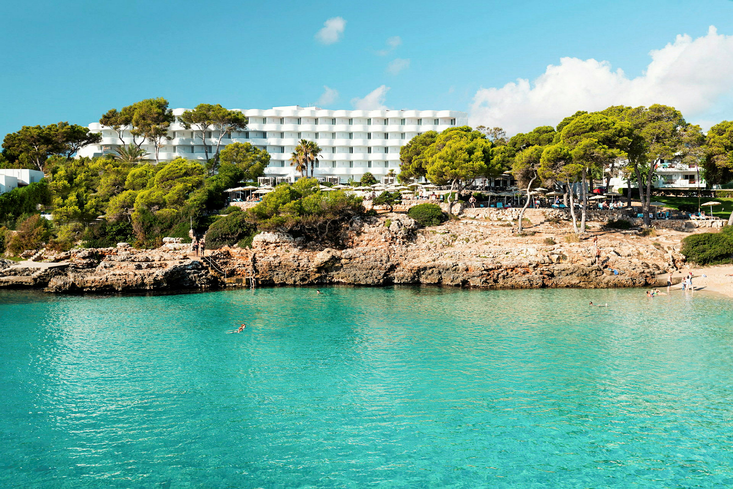 Hotell med hvite bygninger bak en klippefylt strand med klart blått vann. (AI-generert tekst)