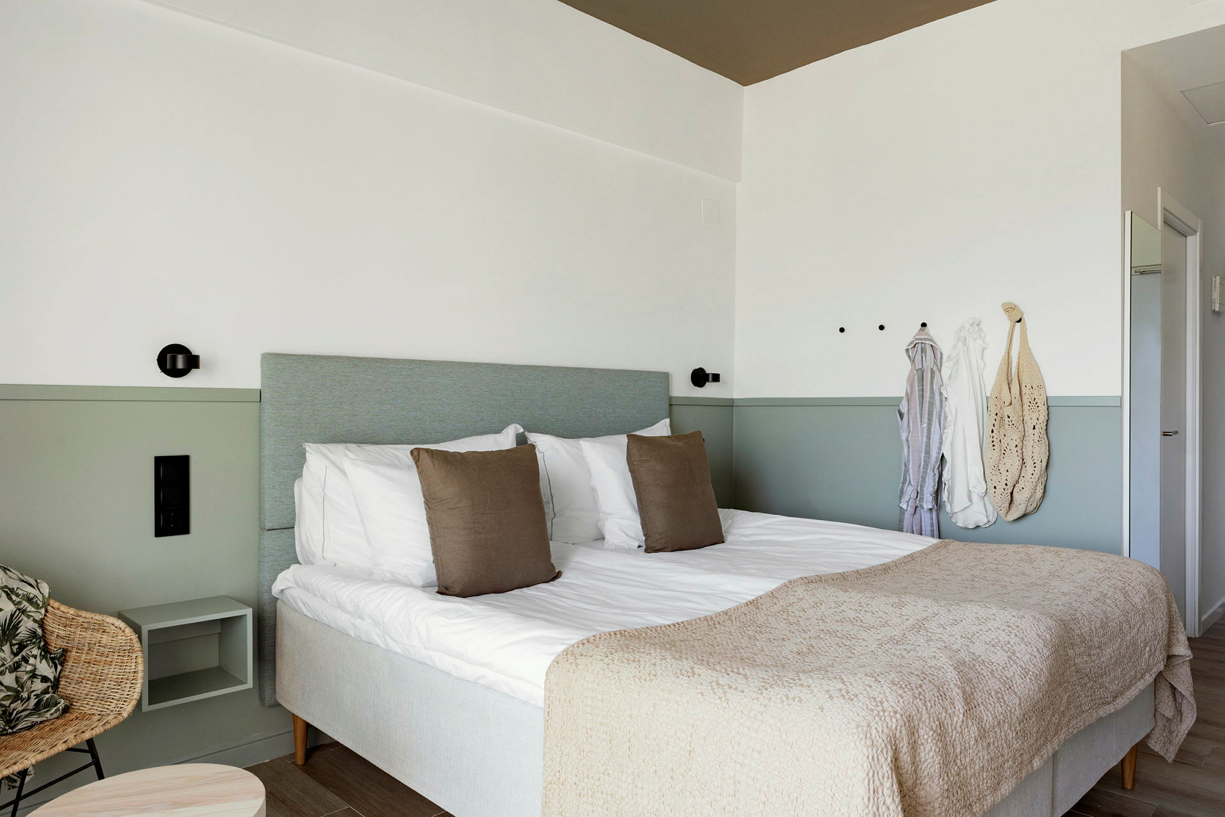 Et hotellrom med en dobbeltseng, to puter og et beige sengeteppe. Vegg med kroker og hengende klær. (AI-generert tekst)