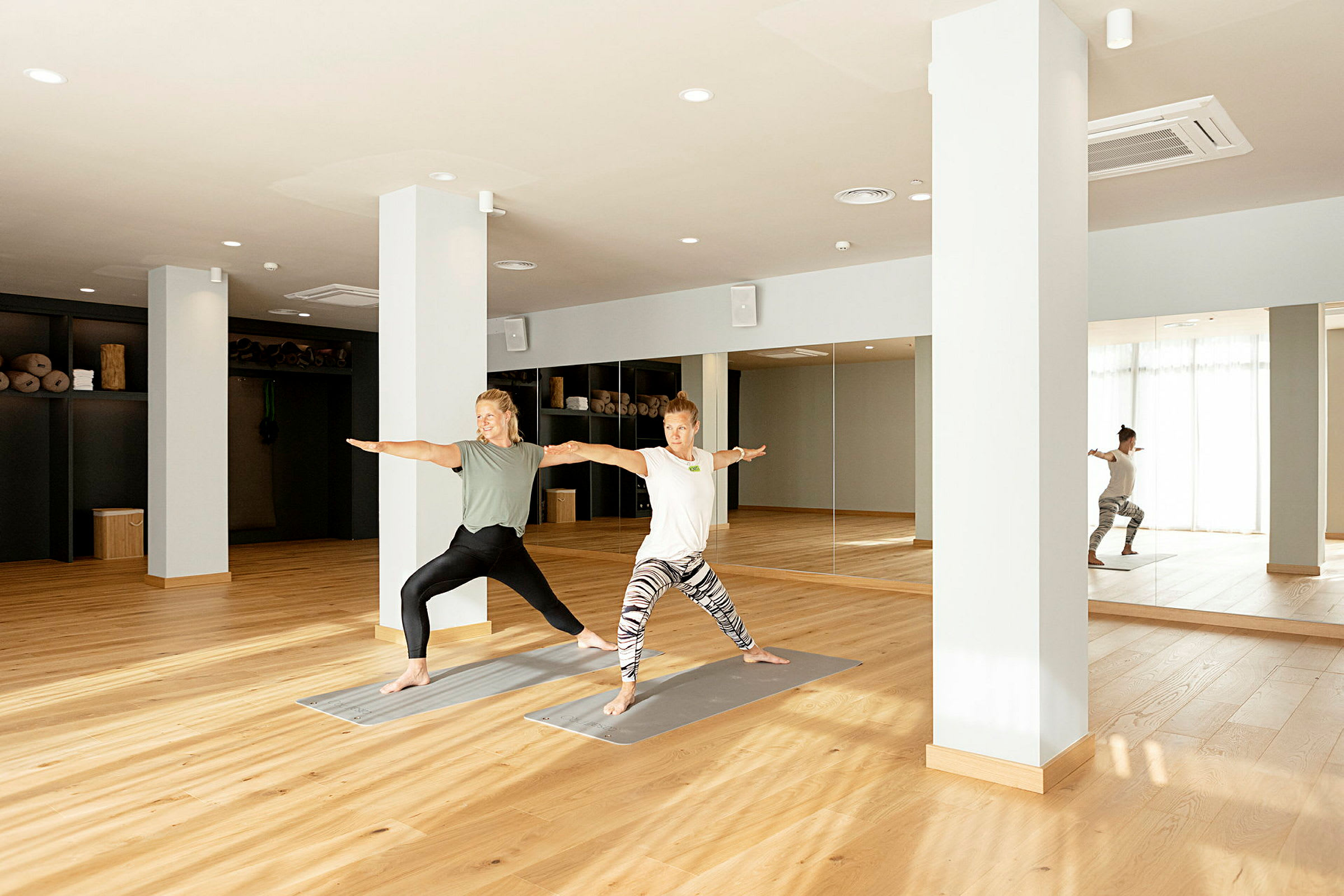 To personer utfører yoga i et lyst studio med tregulv og speilvegg. (AI-generert tekst)