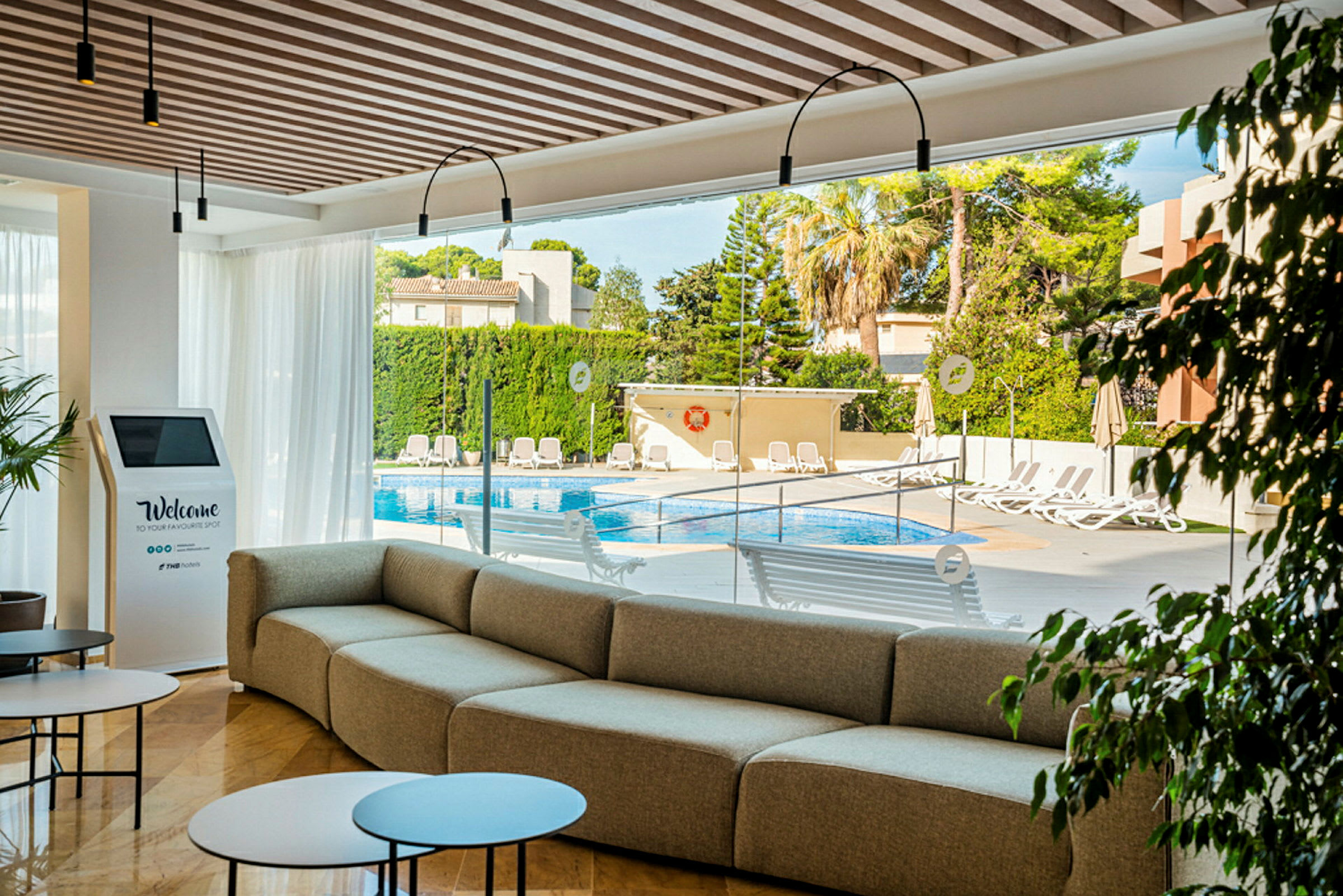 En moderne lounge med sofa, bord og utsikt mot bassengområdet utenfor glassveggene. (AI-generert tekst)