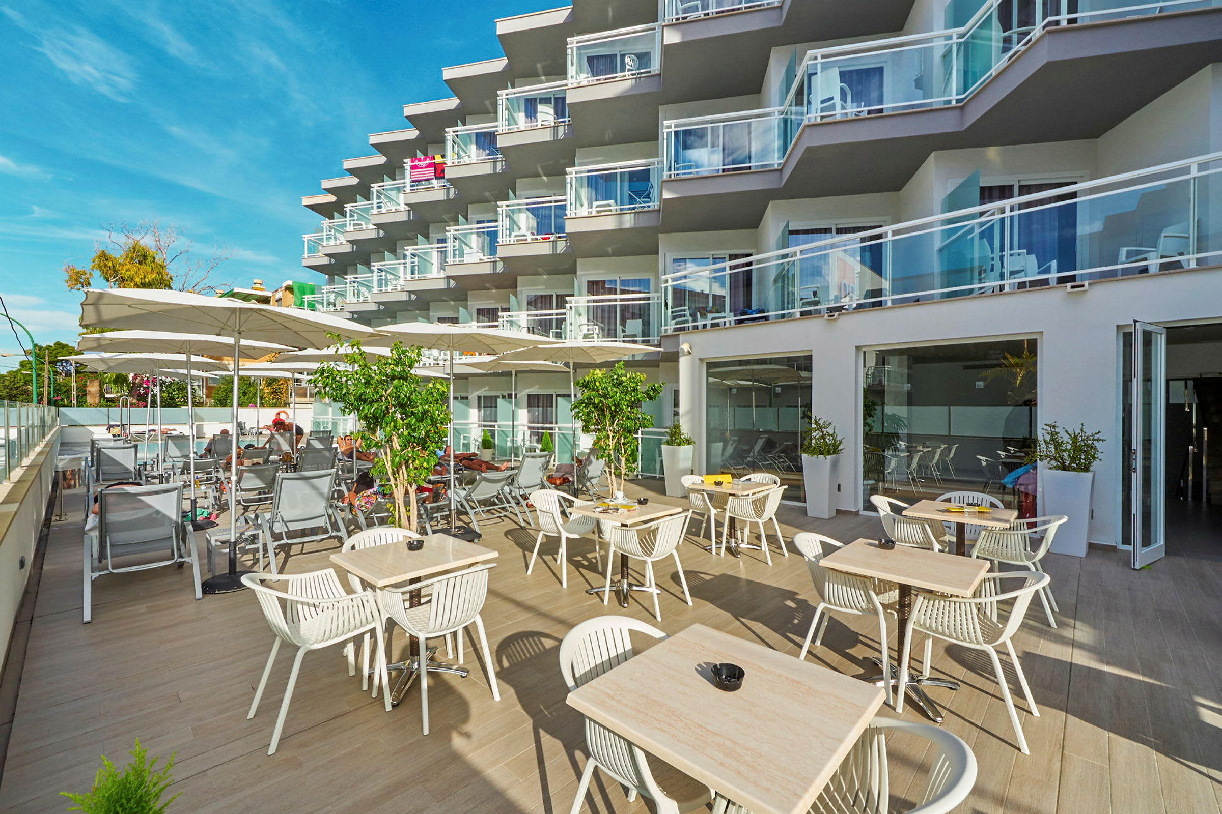 Utendørsområde med bord og stoler samt solsenger ved et hotell med balkonger. (AI-generert tekst)