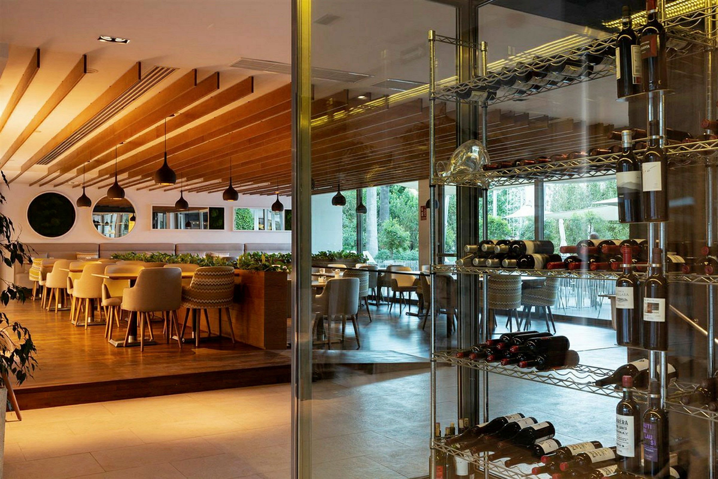 En lys restaurant med tretakdetaljer, store vinduer med utsikt mot grøntområder, og vinflasker på metallhyller. (AI-generert tekst)