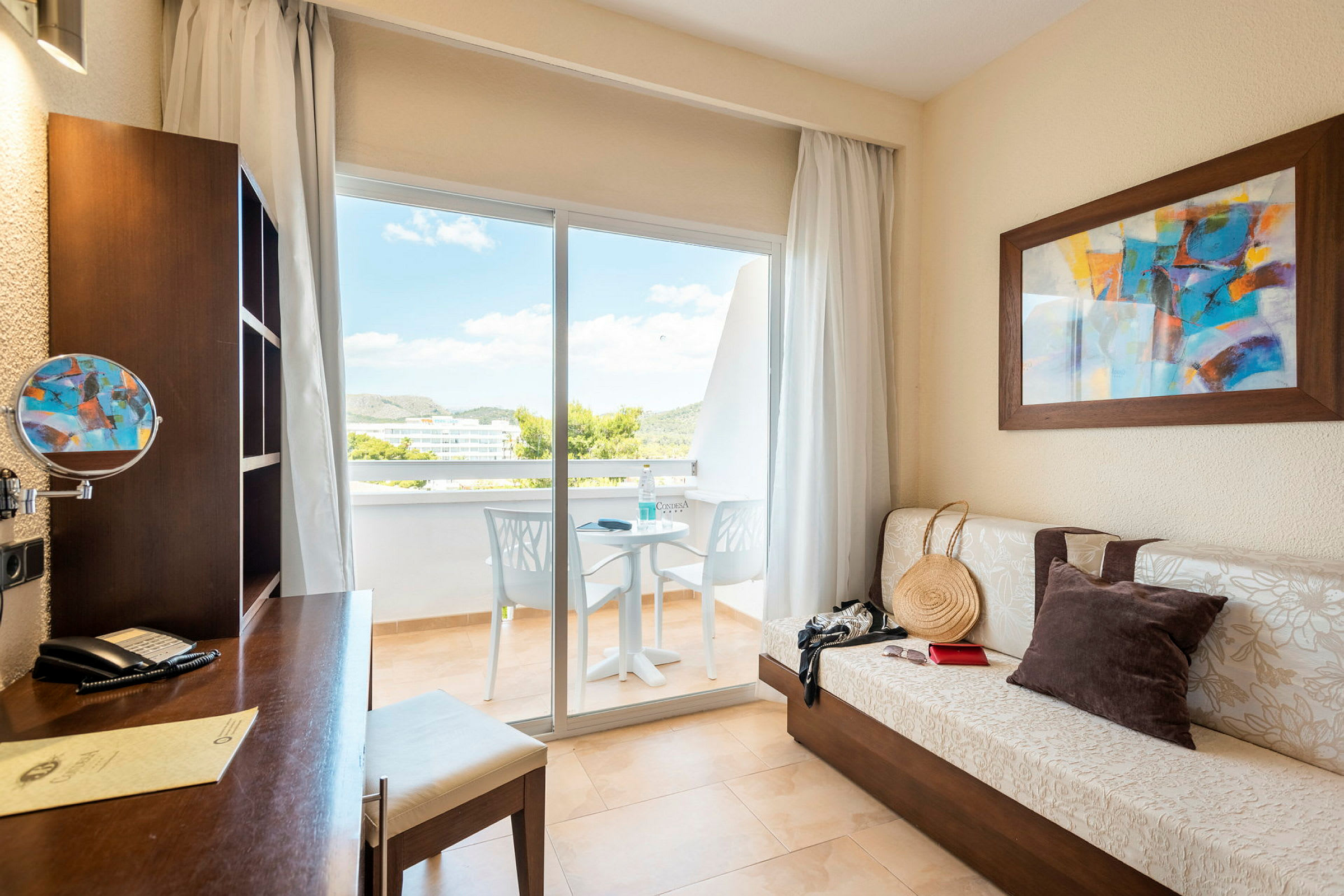 Et hotellrom med skrivebord, sofa, maleri og utgang til balkong. (AI-generert tekst)