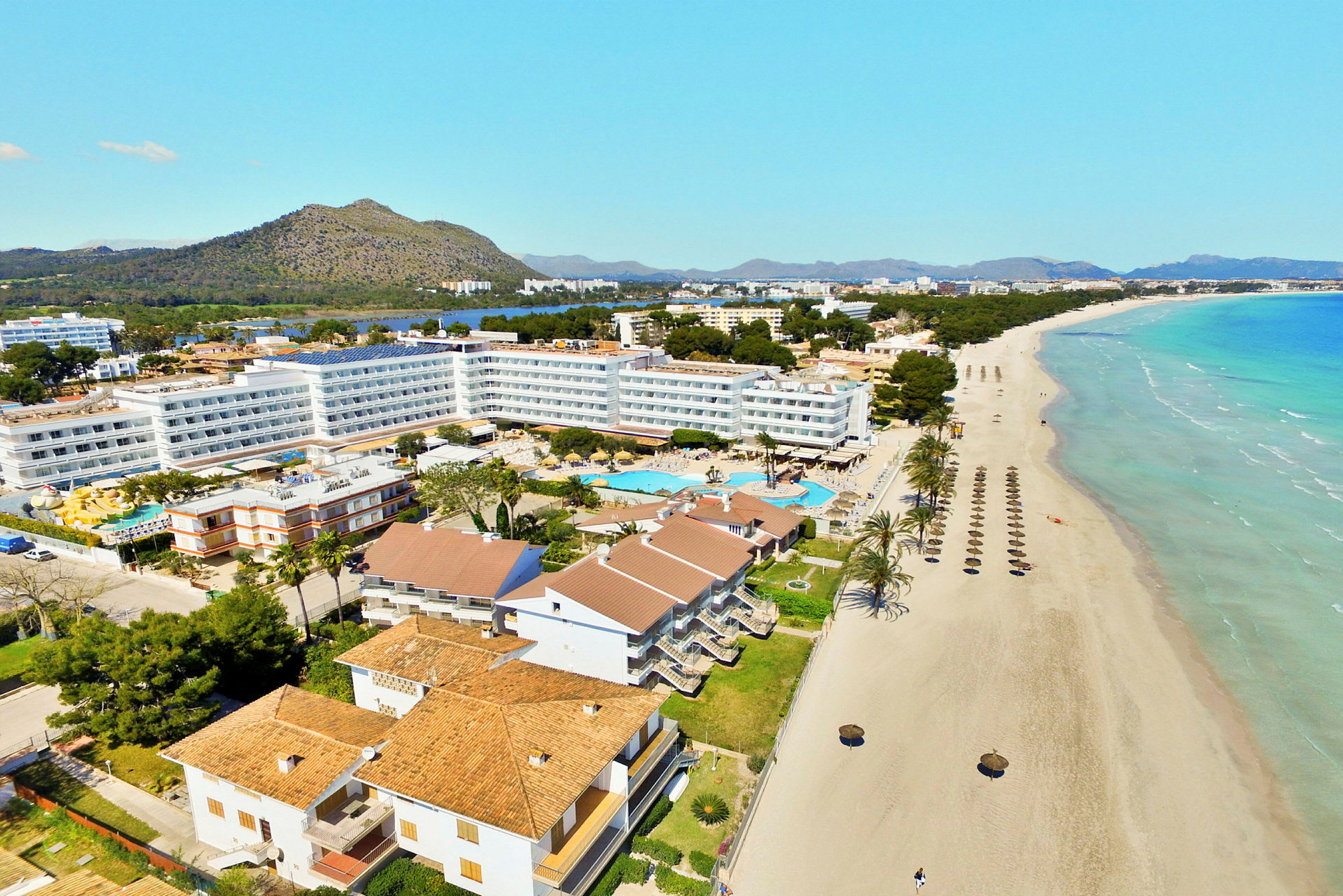 Hotellbygg med bassengområde ved en lang sandstrand med parasoller, samt fjell og vegetasjon i bakgrunnen. (AI-generert tekst)