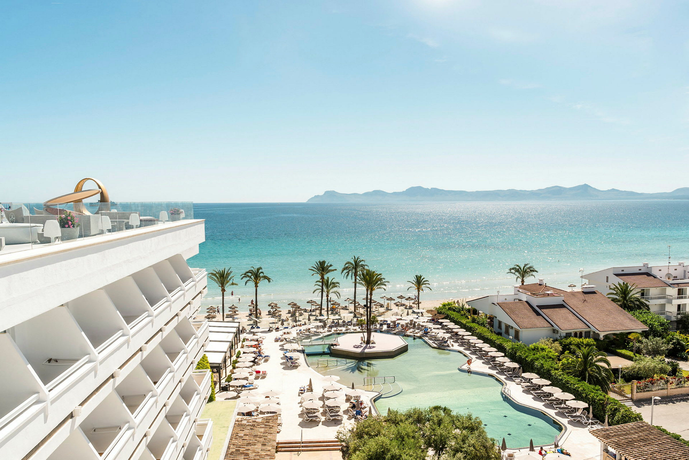 En utsikt over hotellbasseng, strand og hav med palmer og bygninger. (AI-generert tekst)