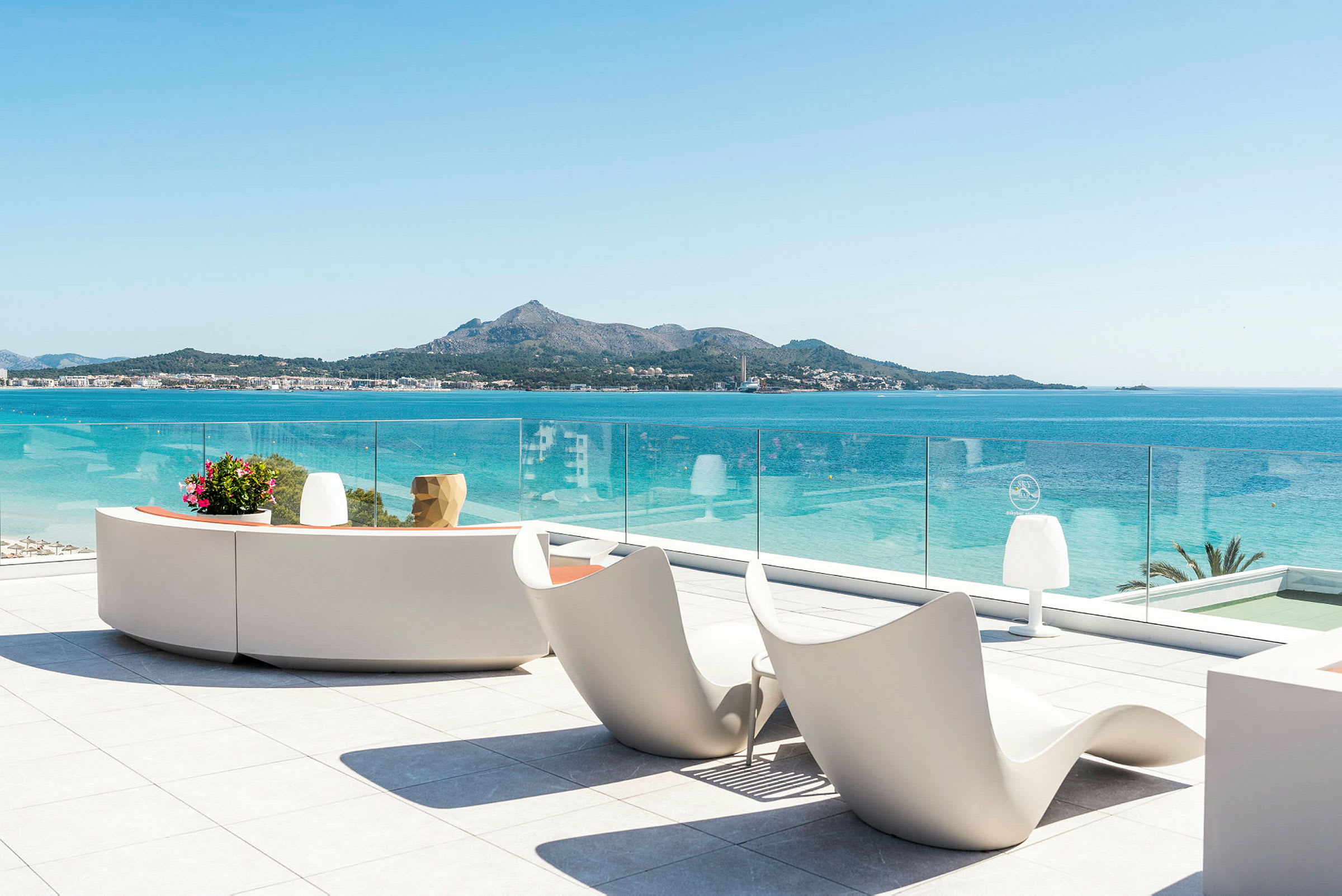 Utsikt fra terrasse med moderne sittemøbler og glassrekkverk mot havet og en fjellkyst. (AI-generert tekst)