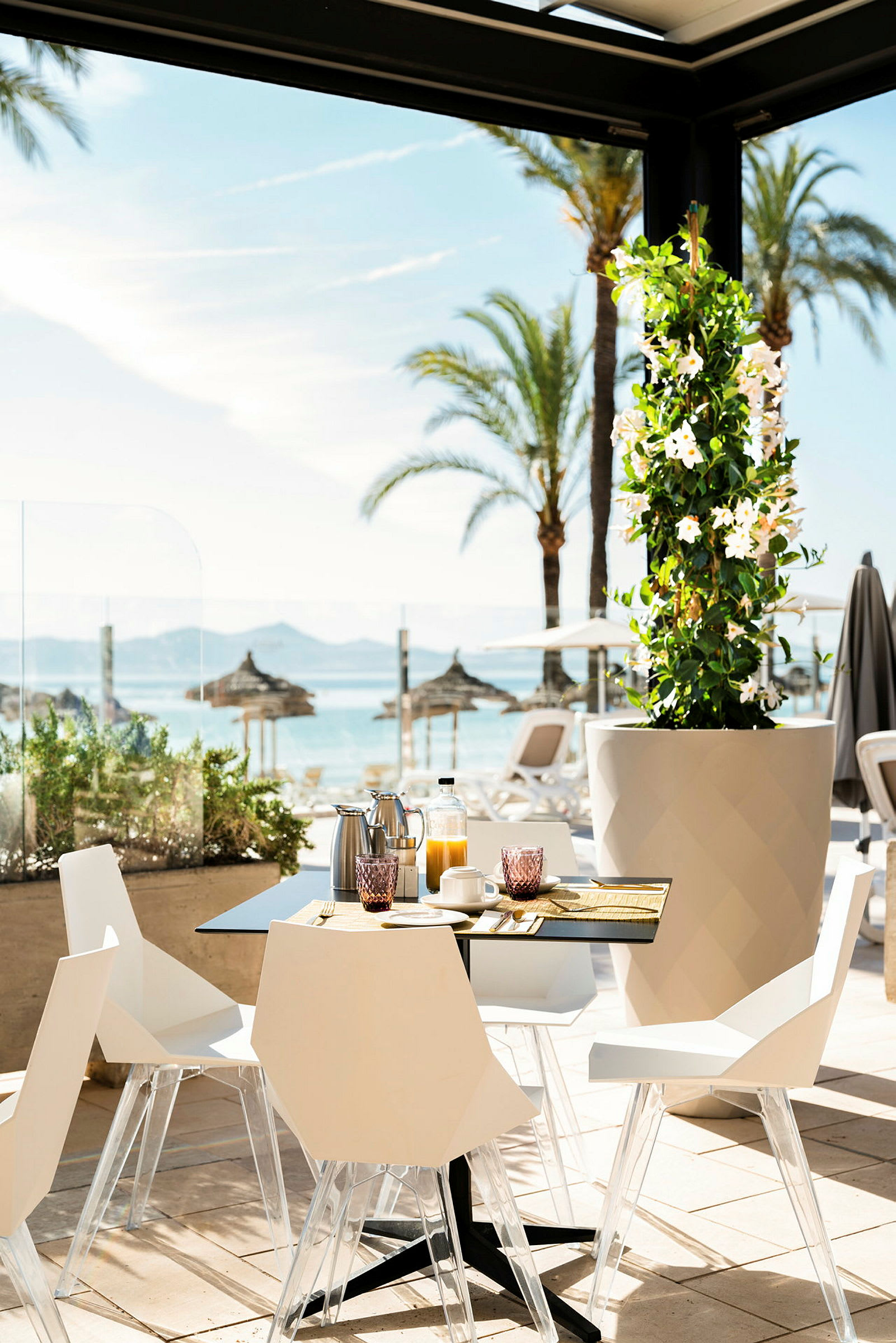 Et bord med stoler, satt opp for frokost, plassert på en solrik terrasse med utsikt over palmer og stranden. (AI-generert tekst)