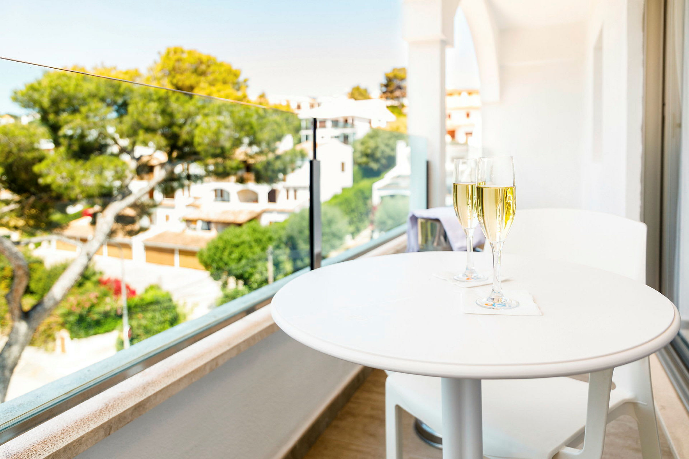 Et hvitt bord med to glass champagne ved en balkong med utsikt over en frodig hage og hus i bakgrunnen. (AI-generert tekst)