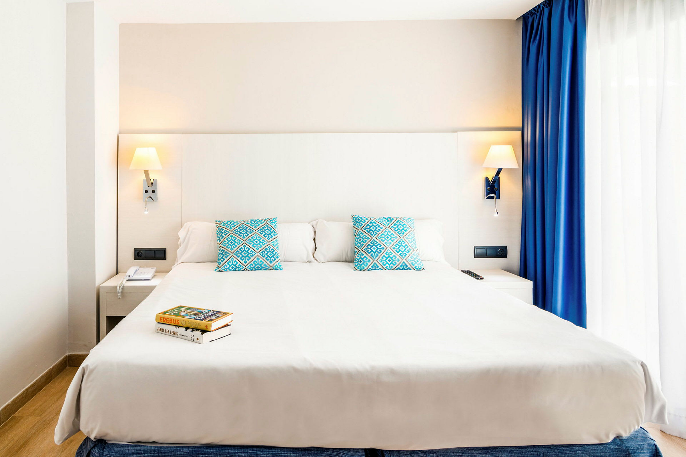 Et moderne hotellrom med en stor dobbeltseng, to blå puter med mønster og leselamper på veggen. Ved siden av sengen står et nattbord og et vindu med hvite og blå gardiner. (AI-generert tekst)