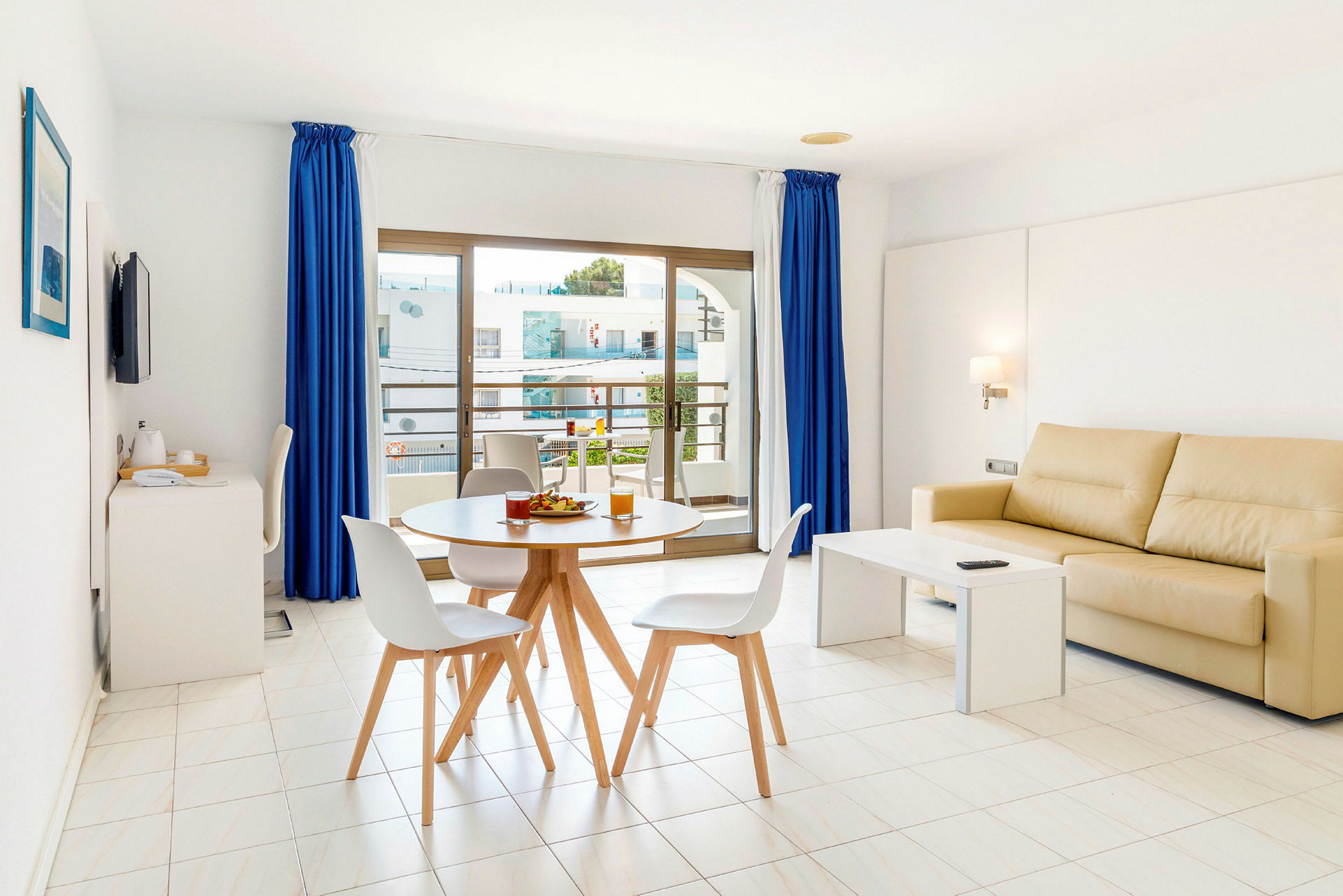 En moderne stue med lys interiør. En beige sofa står ved et hvitt sofabord, og et rundt spisebord med stoler er plassert foran store vinduer med blå gardiner. (AI-generert tekst)