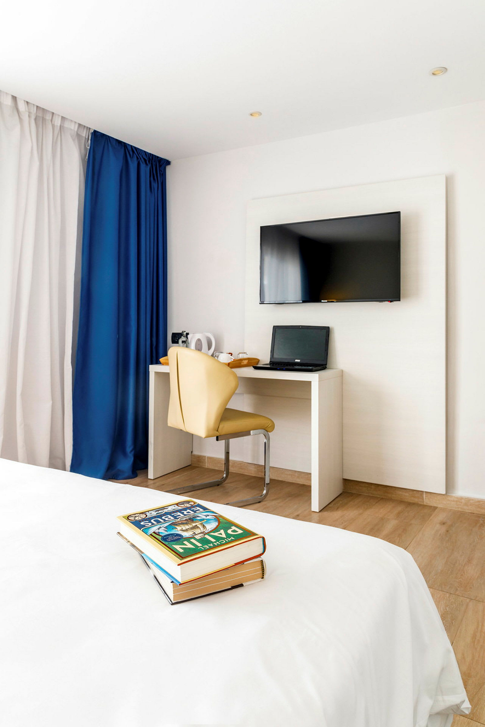 Et hotellrom med en flatskjerm-TV på veggen, et skrivebord med en bærbar PC og en beige stol. I forgrunnen er en seng med en bunke bøker. (AI-generert tekst)