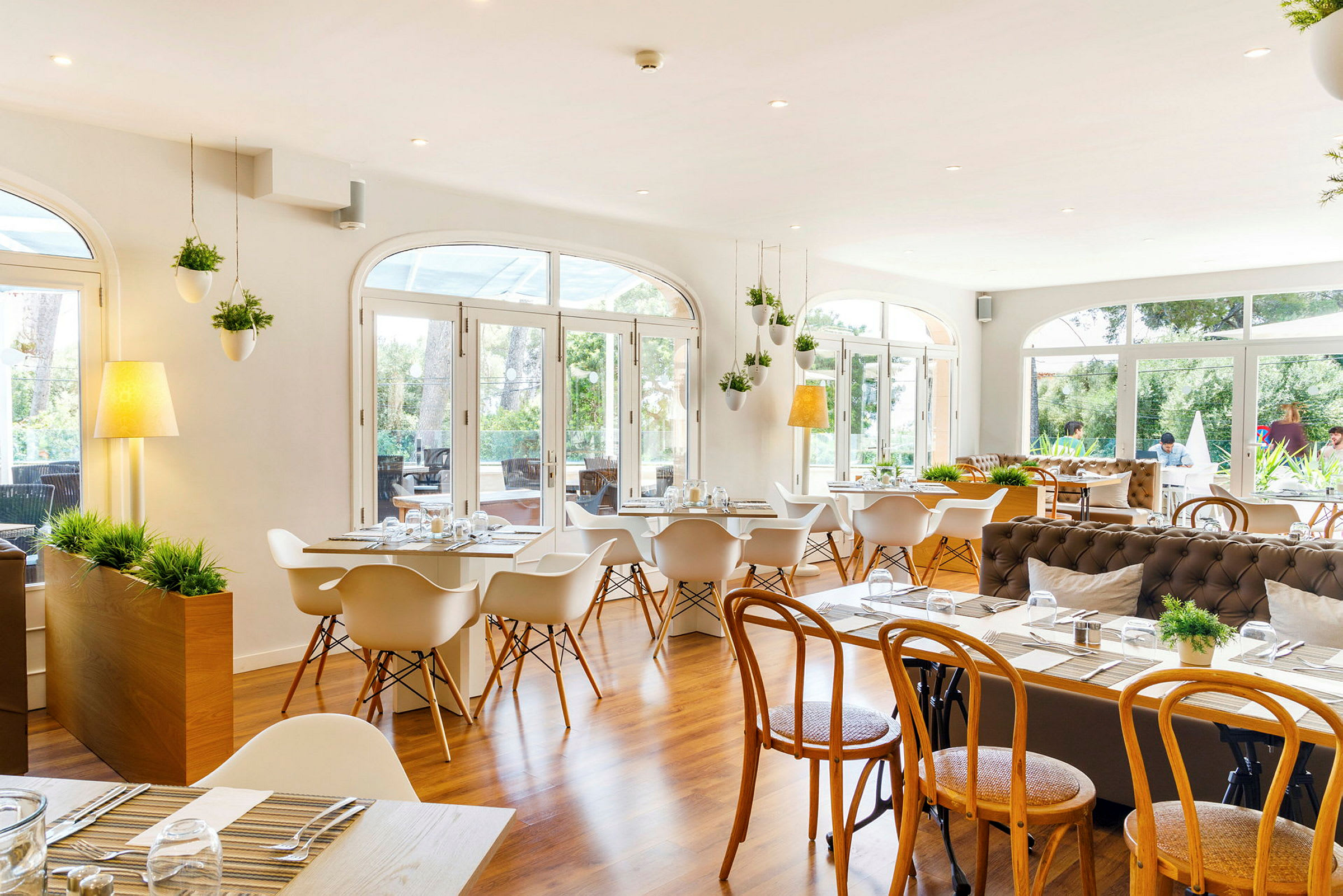 Et lyst og romslig restaurantområde med store vinduer som vender ut mot en grønn utsikt. Rommet er innredet med moderne hvite stoler og tremøbler, med dekorative planter som henger i taket. (AI-generert tekst)