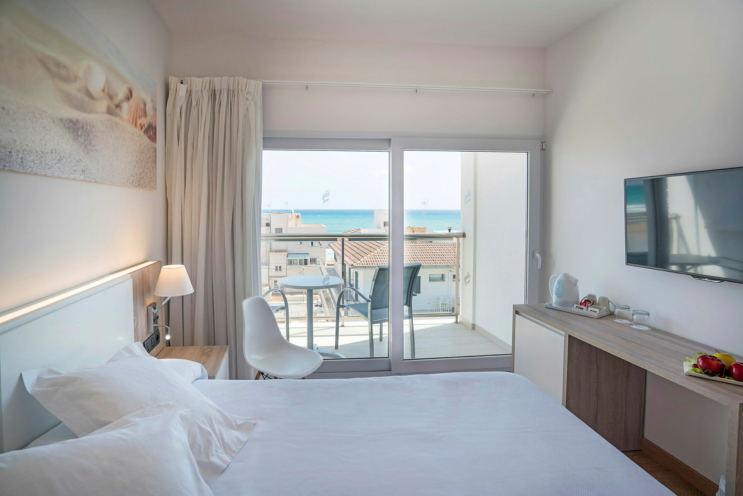 Et hotellrom med seng, skrivebord og en balkong med utsikt mot havet. (AI-generert tekst)