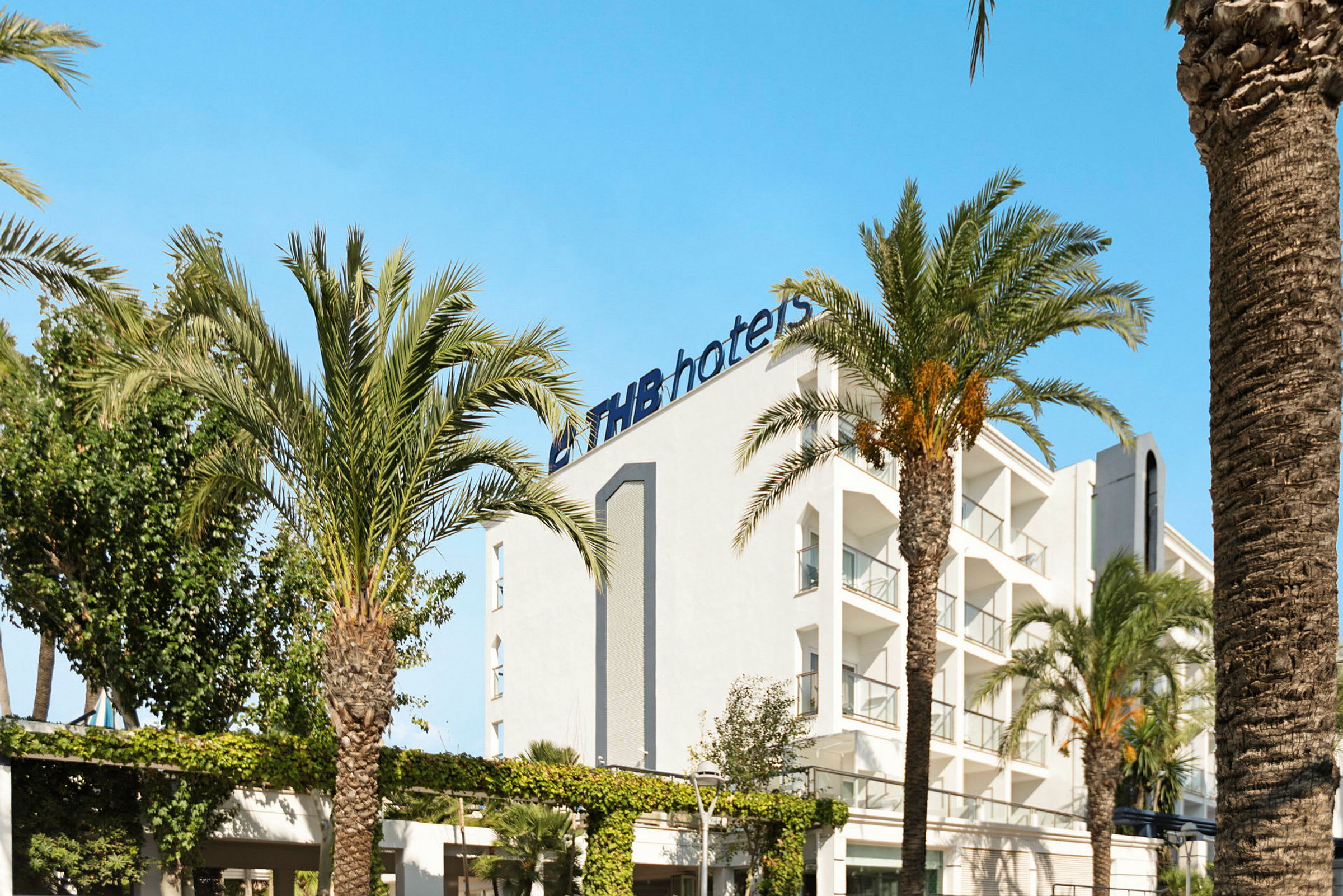 Hvit bygning med logoen 'THB hotels' omgitt av palmer under en klarblå himmel. (AI-generert tekst)