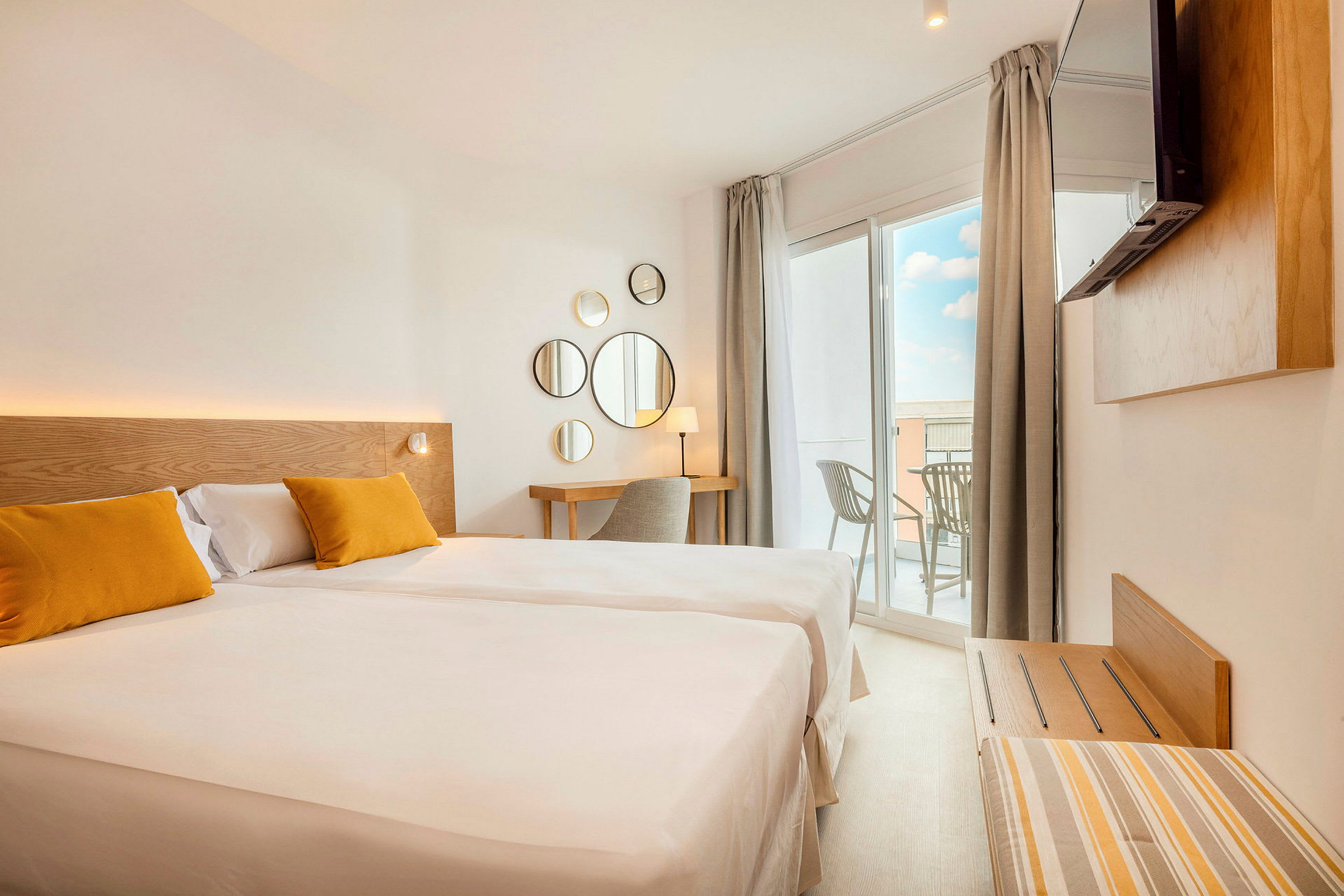 Et lyst hotellrom med en stor seng, to gule puter, en skrivebordshjørne og en balkong med utsikt. (AI-generert tekst)