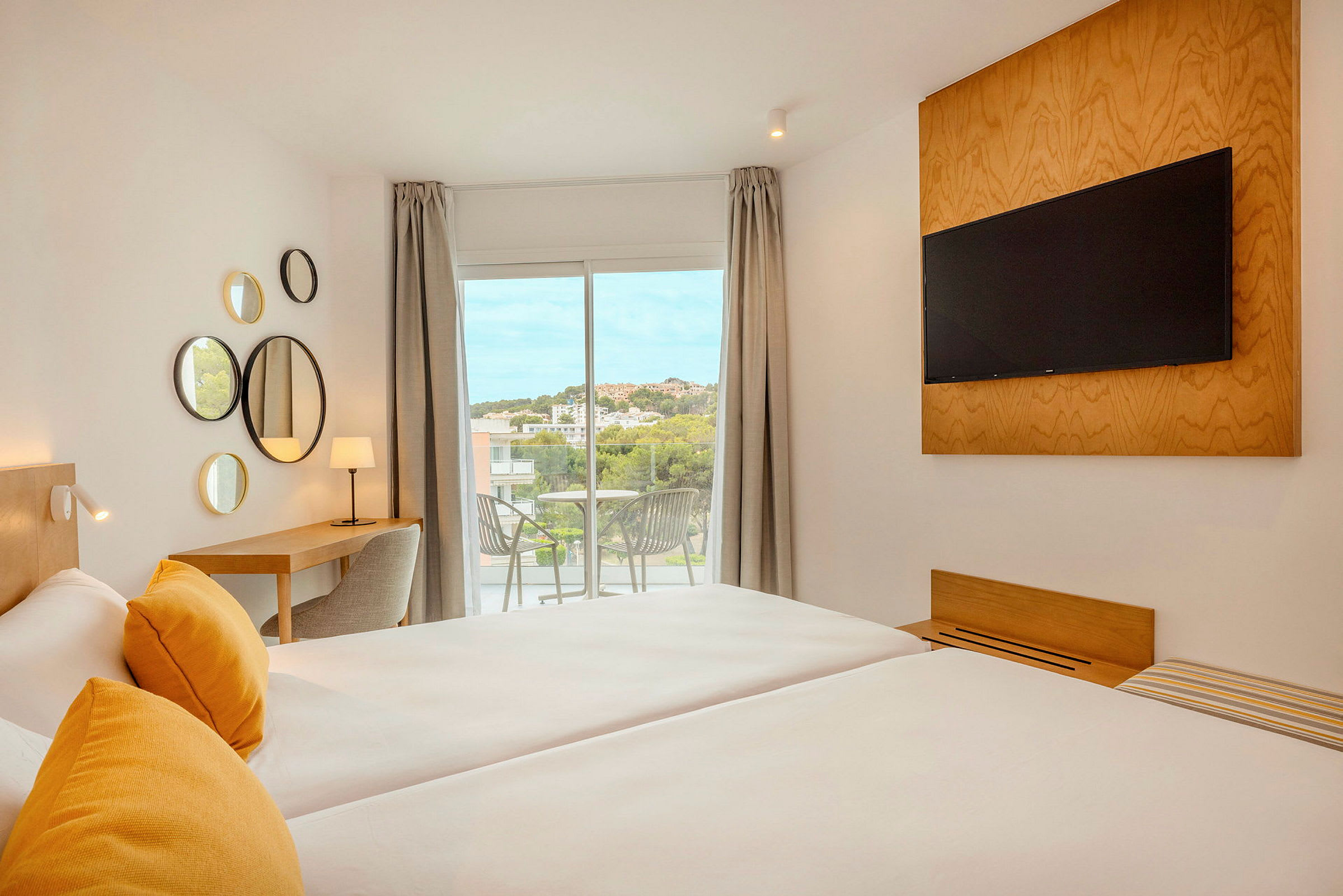 Et moderne hotellrom med to senger, gule puter, en TV montert på veggen, et skrivebord med lampe og en balkong med utsikt. (AI-generert tekst)