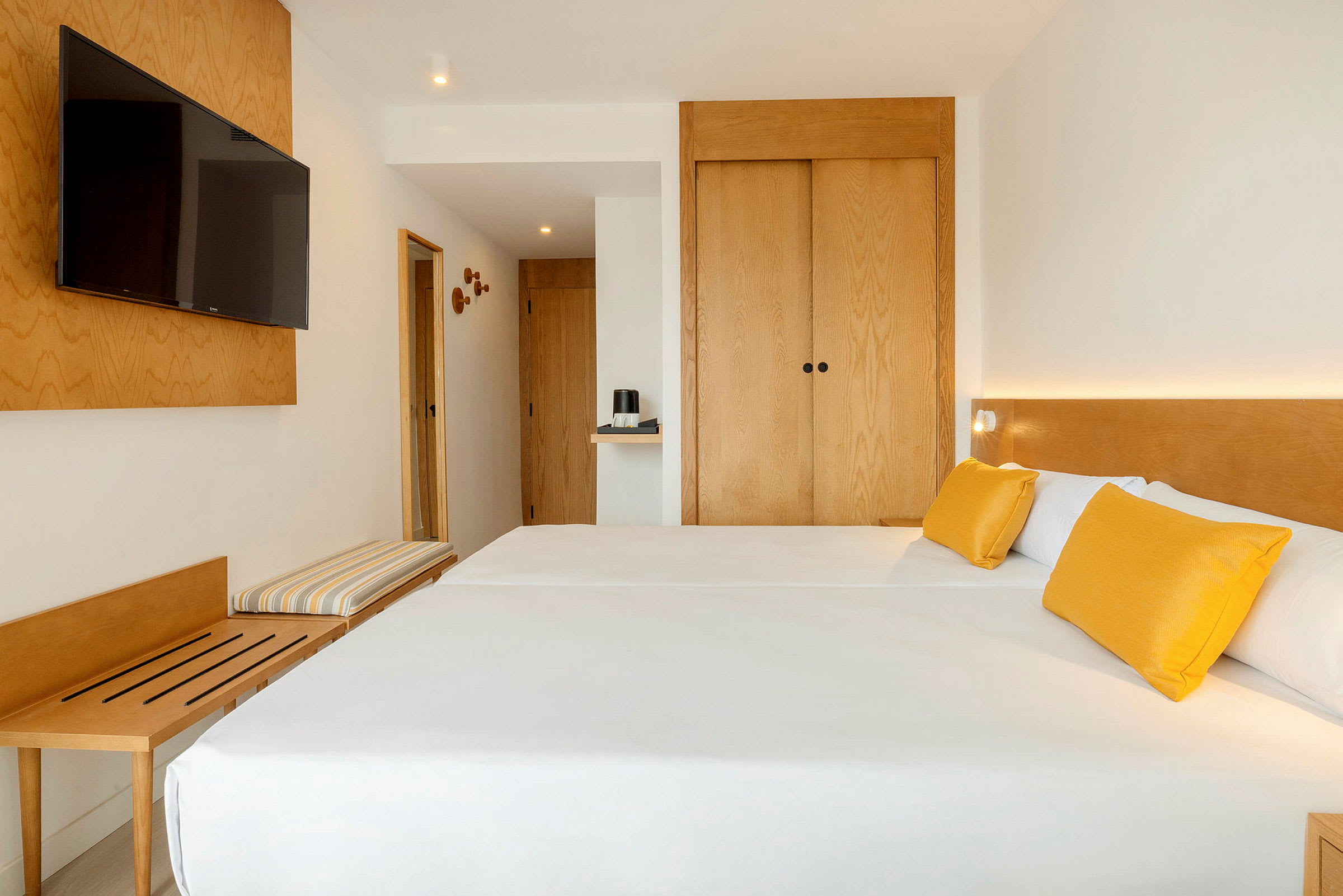 Et hotellrom med moderne innredning, to enkeltsenger med hvitt sengetøy og gule puter, en veggmontert TV og tremøbler. (AI-generert tekst)