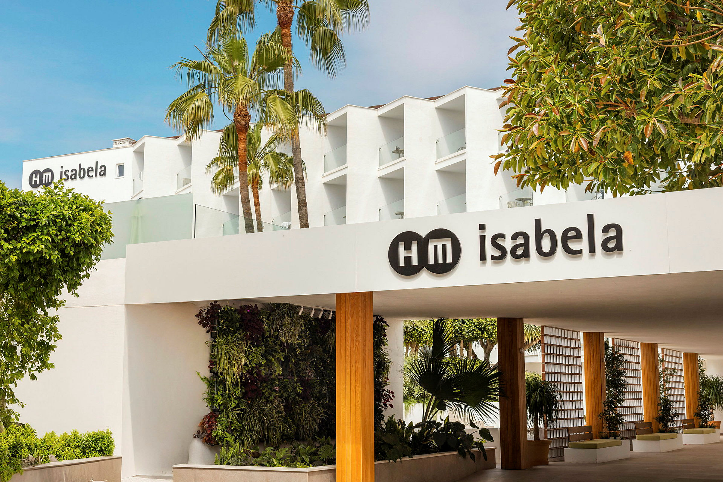Bilde av en hotellbygning med navnet "HM Isabela" synlig på skiltet. Palmetrær og grøntområder er i omgivelsene. (AI-generert tekst)