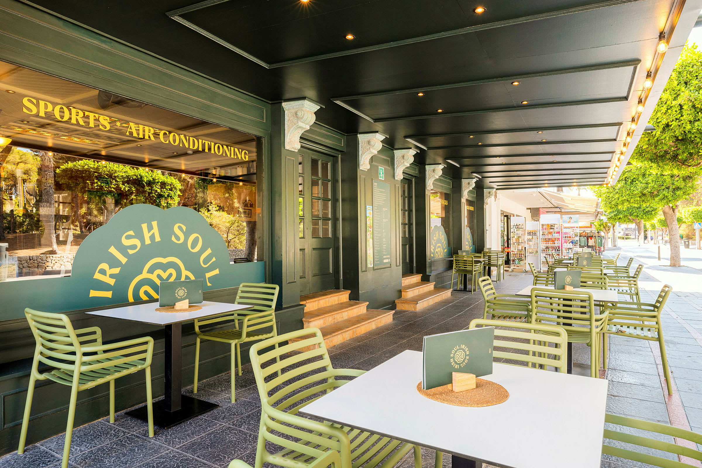 Uteservering med grønne stoler og hvite bord foran en restaurant med grønn fasade. (AI-generert tekst)
