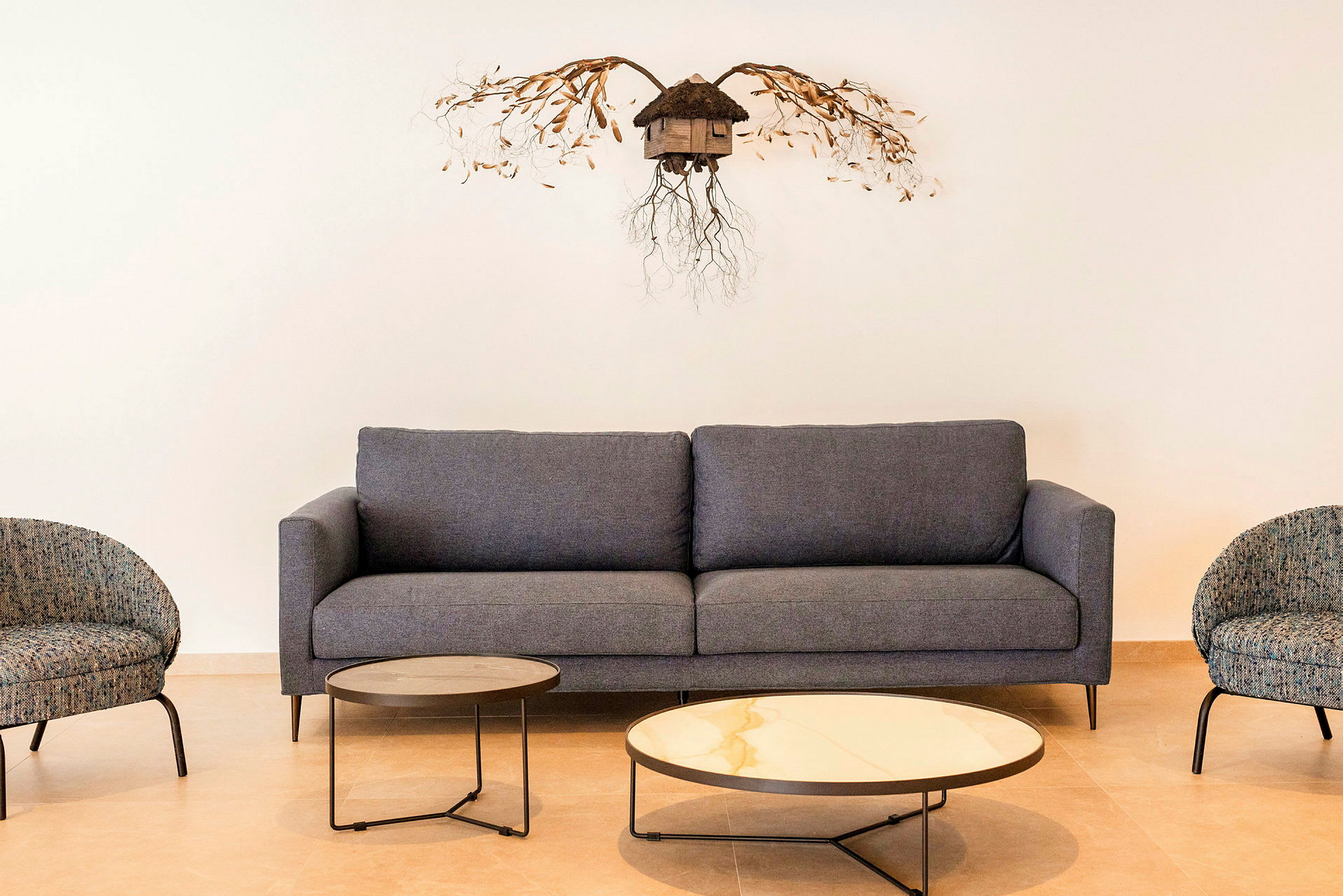 En stilren grå sofa med to runde kaffebord foran og to lenestoler ved siden. På veggen henger en dekorasjon. (AI-generert tekst)