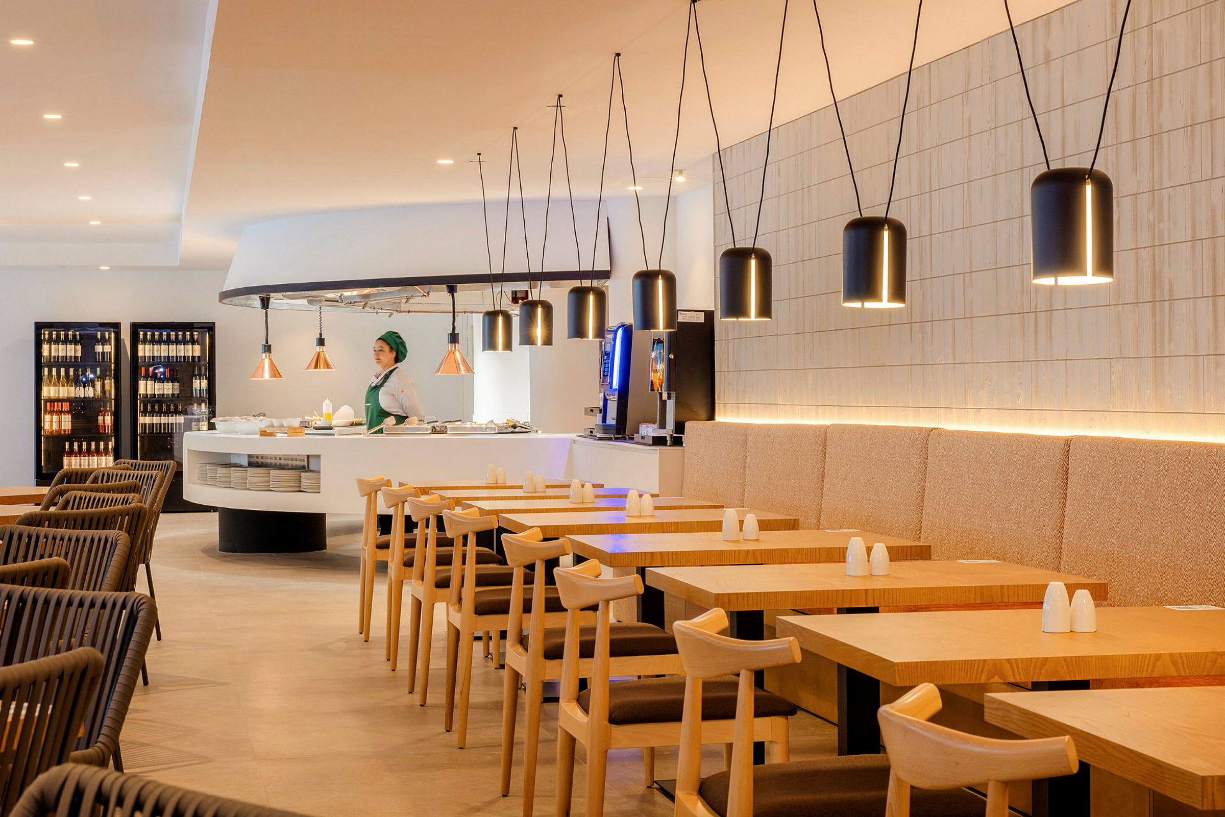 Bilde av et moderne restaurantmiljø med trebord og stoler. En person står ved en serveringsdisk omgitt av hengende lamper. (AI-generert tekst)