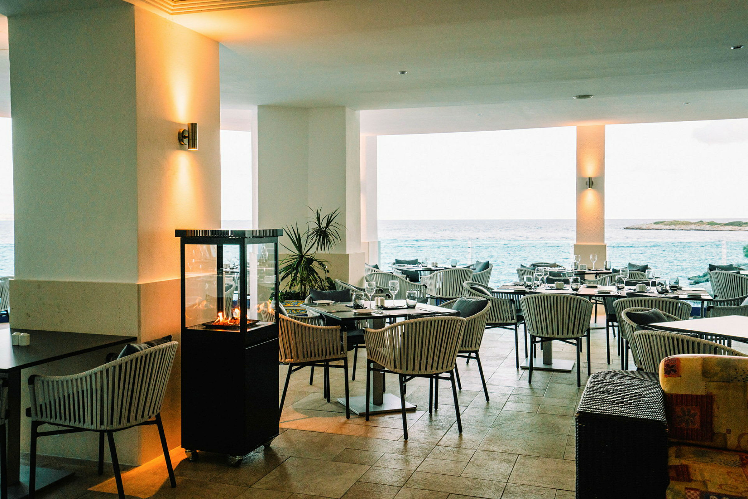 En restaurant med utendørsutsikt over havet, møblert med bord og stoler og dekorert med en liten peis. (AI-generert tekst)