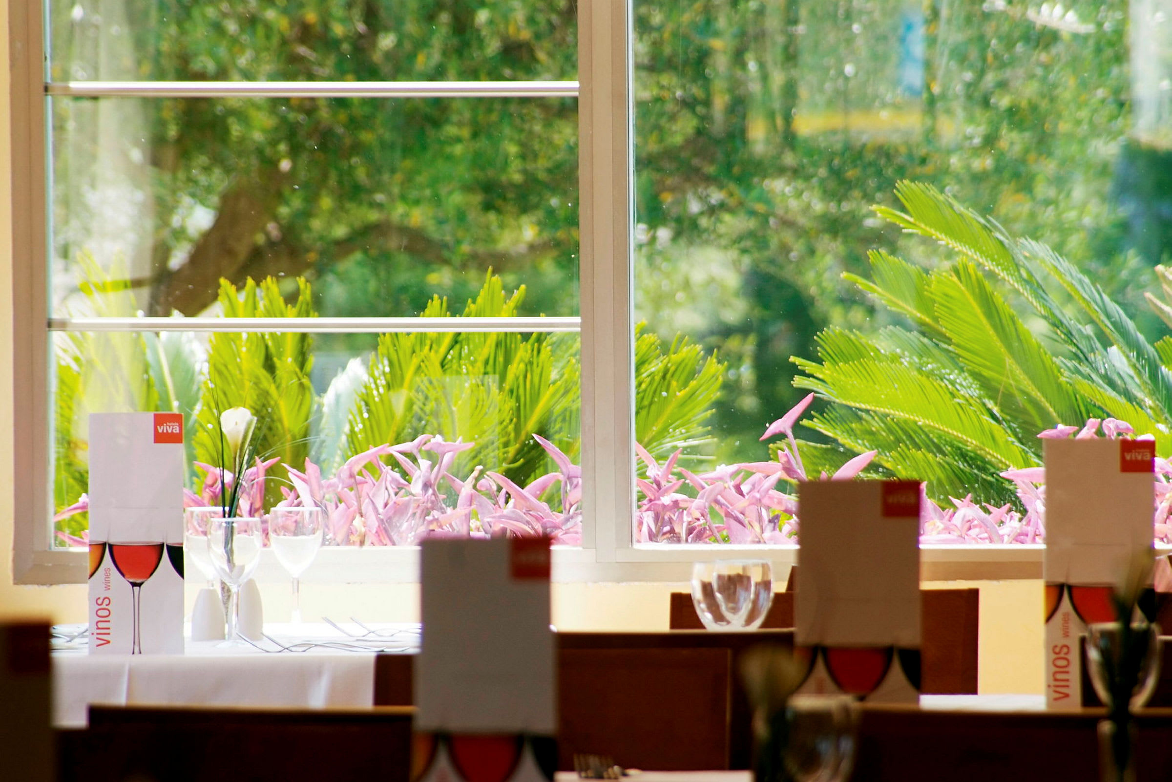 Et restaurantbord dekket med menyer, glass og bestikk foran et vindu som vender ut mot en frodig hage. (AI-generert tekst)