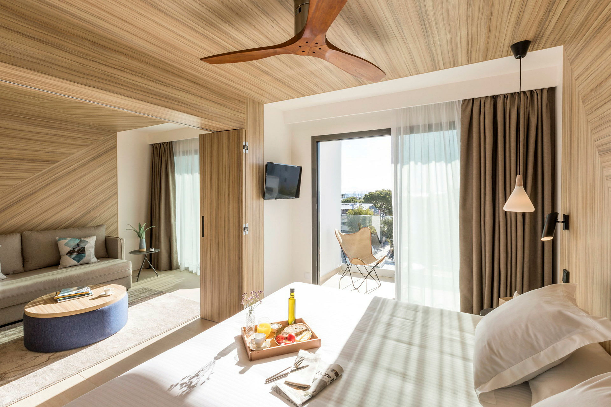 Et moderne hotellrom med trematerialer og store vinduer som slipper inn naturlig lys. Det finnes en seng med en frokostbrett, en sofa og en balkong med utsikt. (AI-generert tekst)