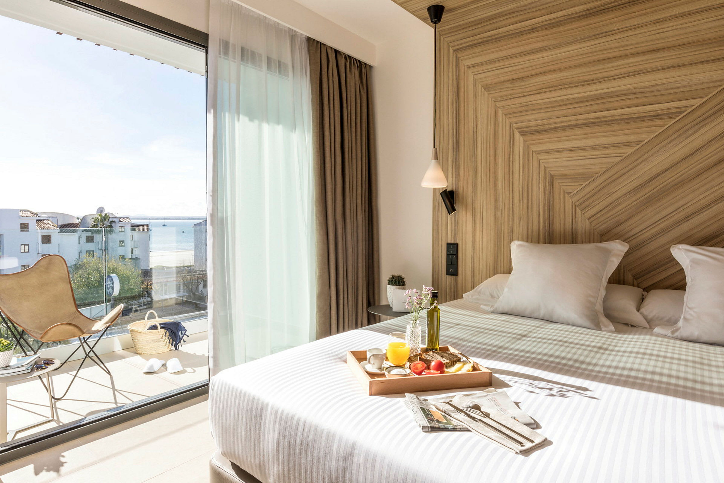 Et lyst hotellrom med en oppredd seng med en frokostbrett. Store glassdører fører ut til en balkong med utsikt over havet. (AI-generert tekst)