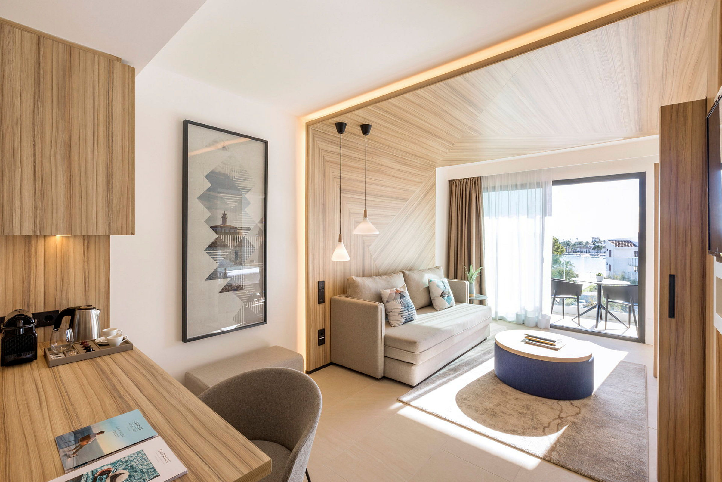 Et moderne hotellrom med lyse tremøbler, inkludert et skrivebord og en sofa, samt et stort vindu som vender ut mot en møblert balkong. (AI-generert tekst)