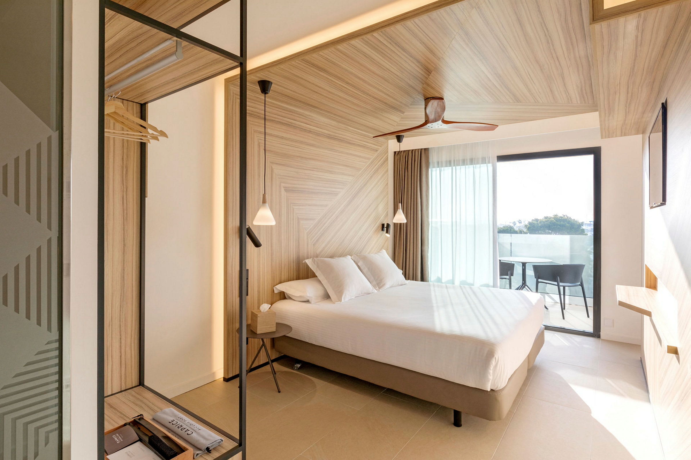 Et hotellrom med en lys og moderne design, en seng med hvitt sengetøy, tremøbler og et vindu som fører til en balkong. (AI-generert tekst)