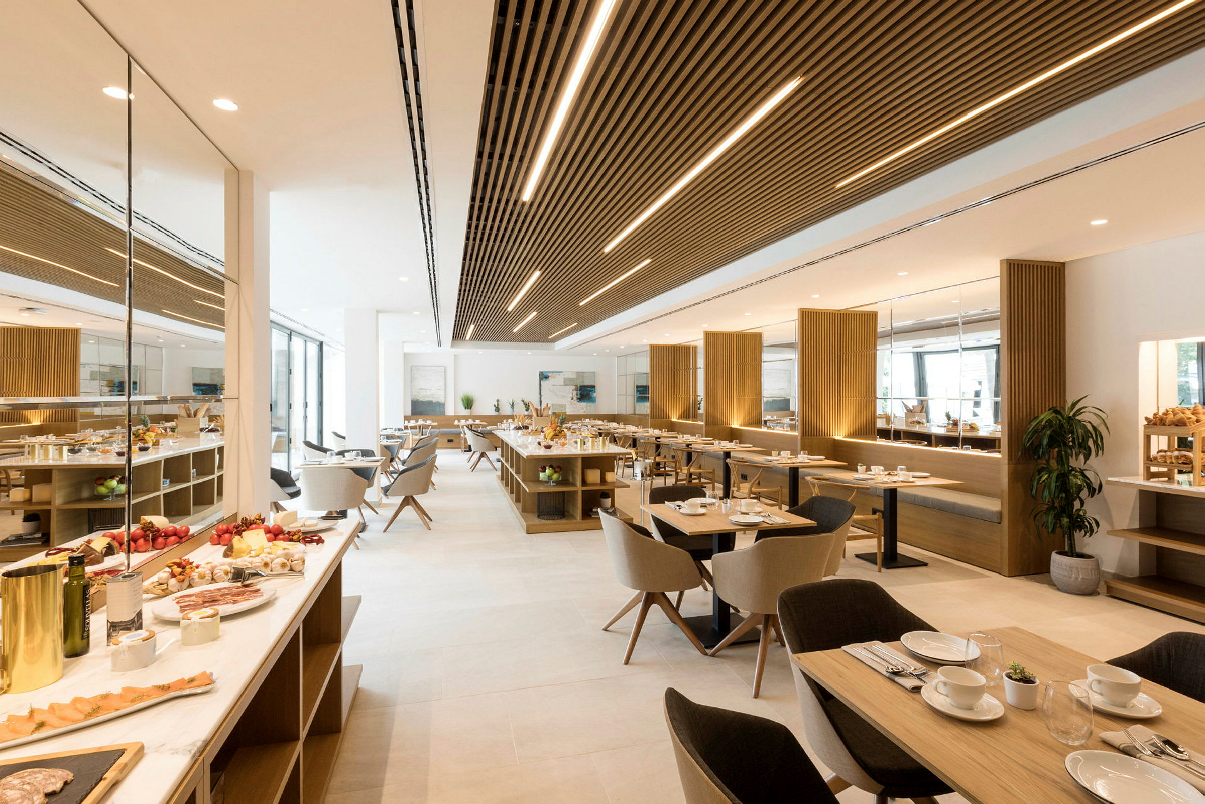 En moderne restaurant med lyst interiør som viser spisebord, spisestoler og serveringsbord. Tallerkener og glass er klare for servering, og fersk mat er framme. (AI-generert tekst)