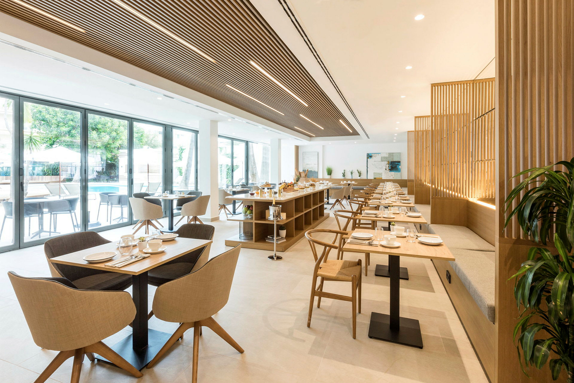 En lys restaurant med bord og stoler i tre og tekstil. Glassvinduer vender ut mot et utendørs basseng. (AI-generert tekst)
