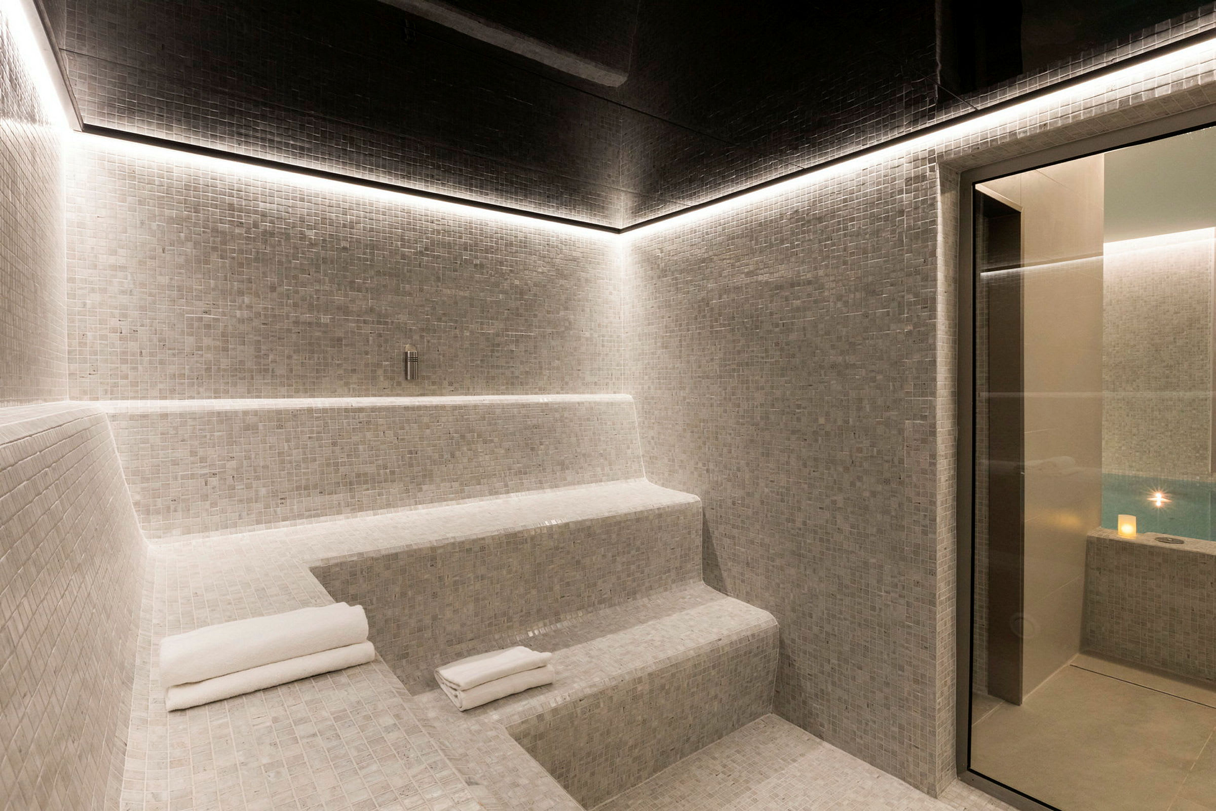 Bilde av et moderne hamam med mosaikkledde sitteplasser og en glasskant som fører til et basseng. (AI-generert tekst)