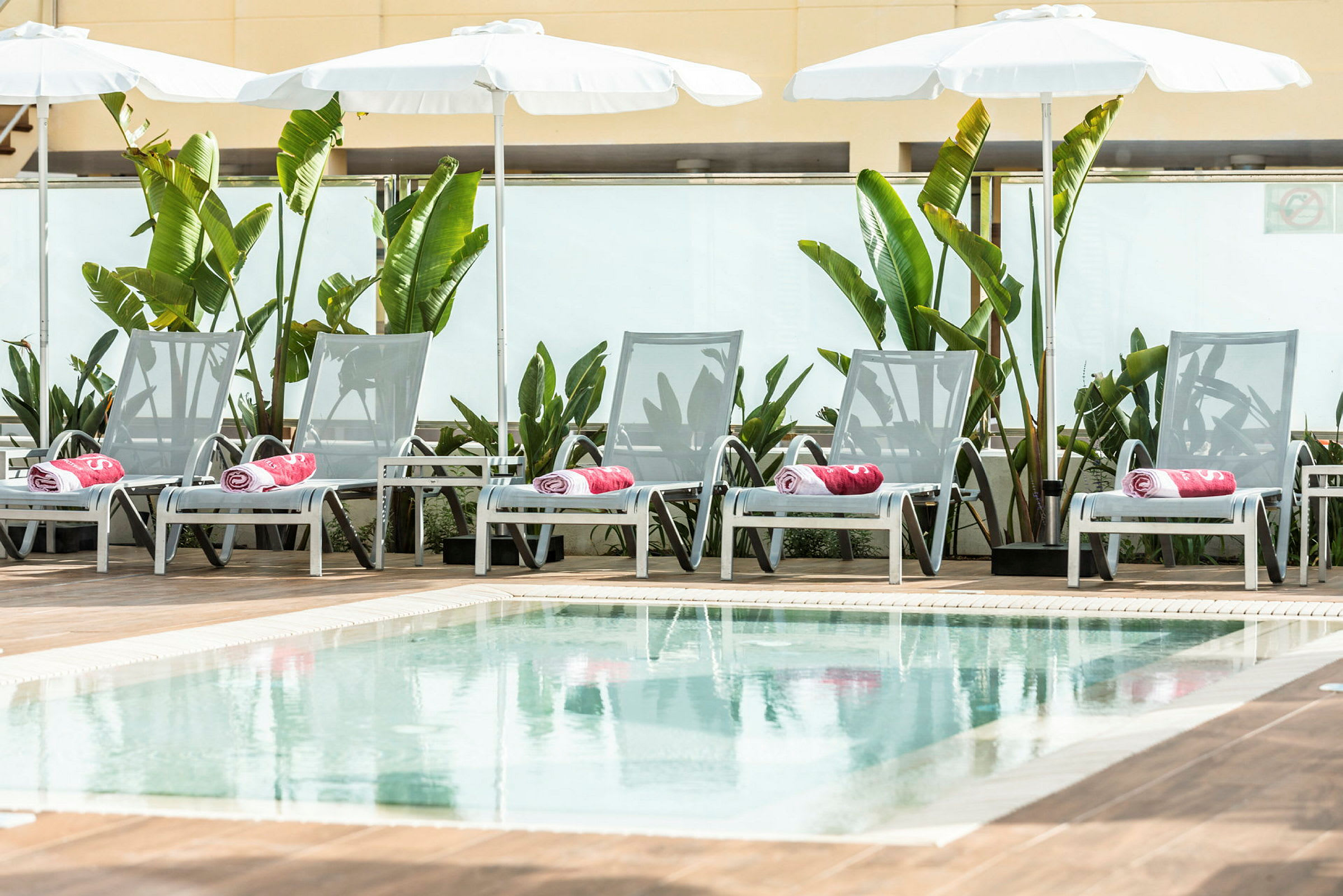 Solstoler ved et basseng, omgitt av grønne planter og parasoller. (AI-generert tekst)