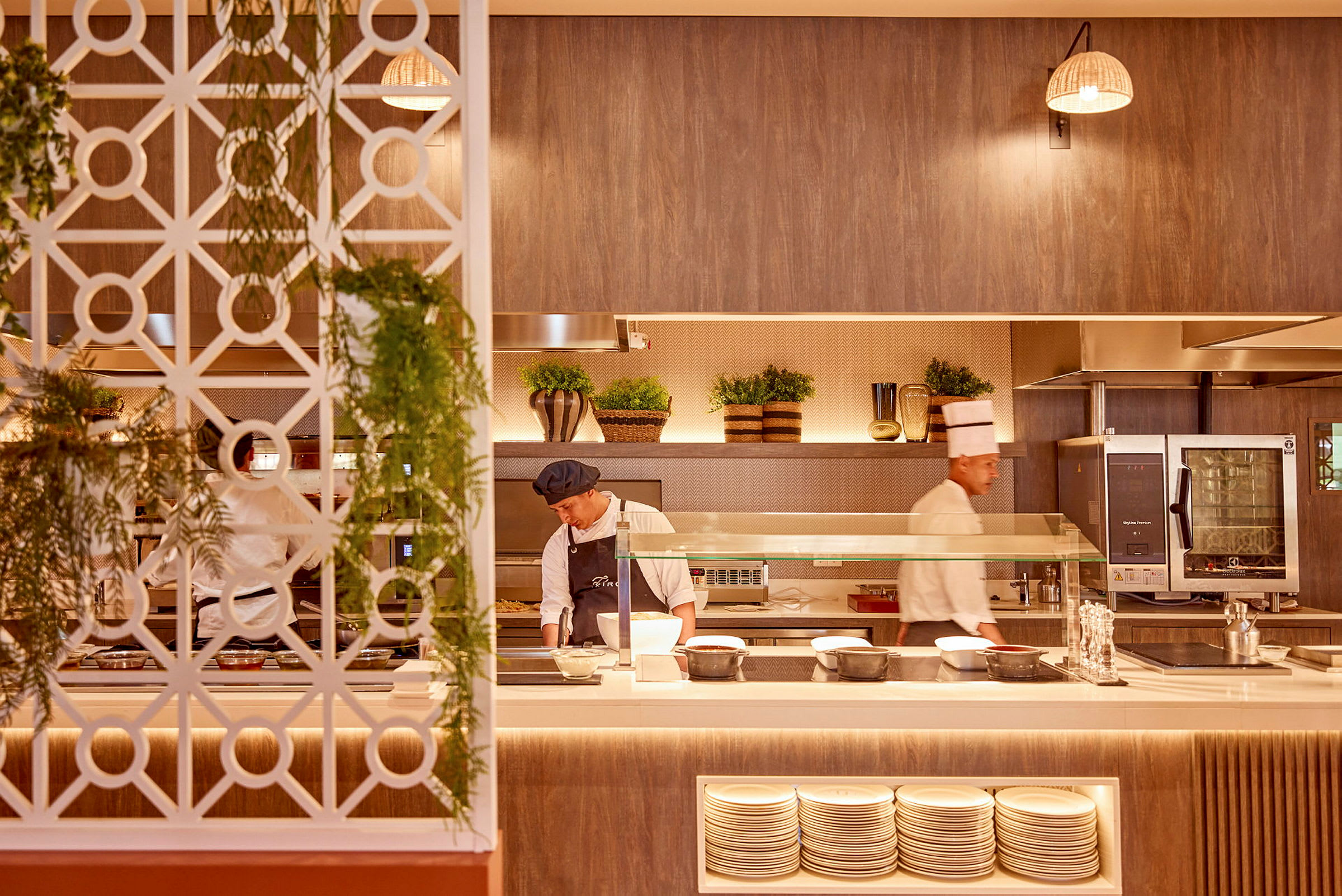 Et restaurantkjøkken med kokker som arbeider, en dekorativ vegg med hengende planter, tallerkener i hyller og interiør med rustikke detaljer. (AI-generert tekst)