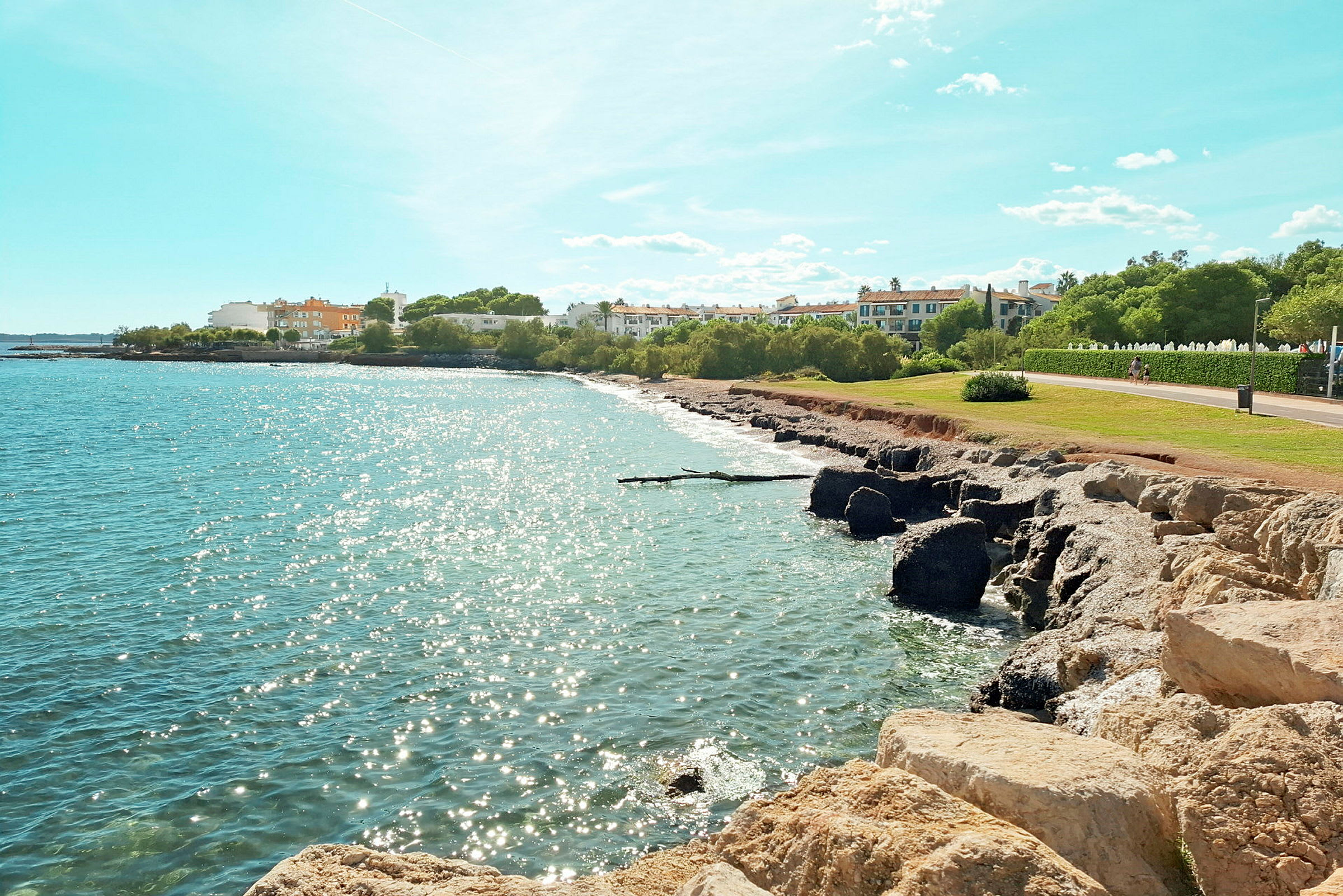 En sandstrand med klipper ved en glitrende kyst, med bygninger og grønne områder i bakgrunnen under en klar blå himmel. (AI-generert tekst)