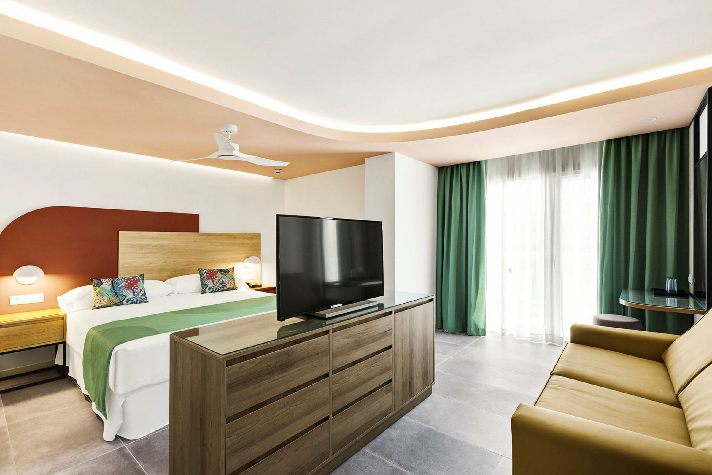 Et hotellrom med en stor seng, en kommode med TV og en sofa samt gardiner foran vinduet. (AI-generert tekst)