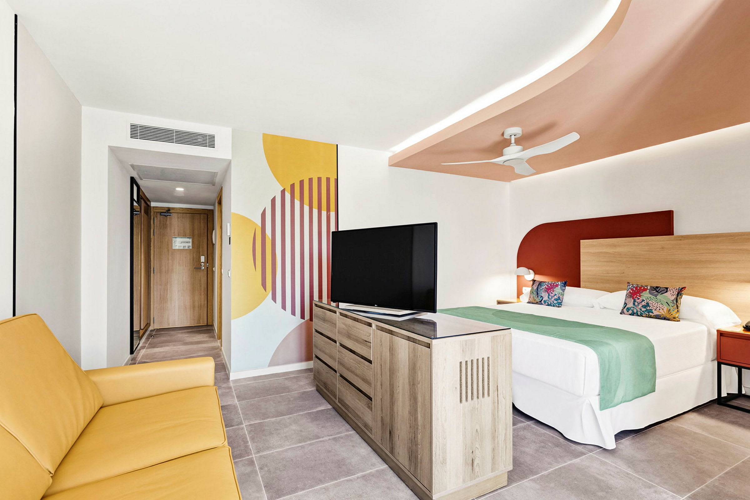 Hotellrom med en seng, gul sofa, TV og dekorativ veggmaleri. (AI-generert tekst)